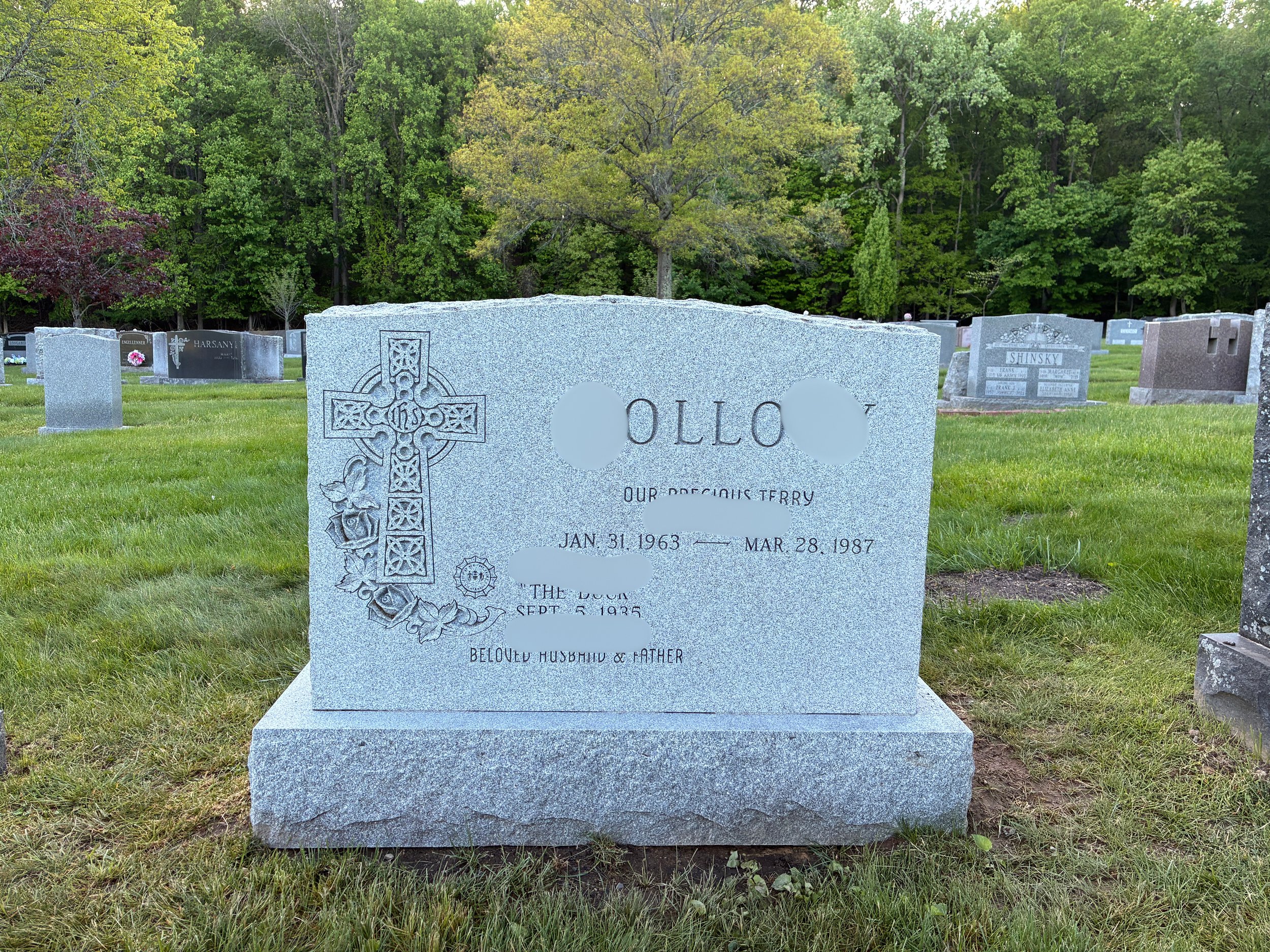 A light gray granite gravestone in a cemetery, marked with a Celtic cross and roses, with inscriptions for Ollico, a person born January 31, 1963, and died March 28, 1987, surrounded by other gravestones and green grass, with trees in the background.