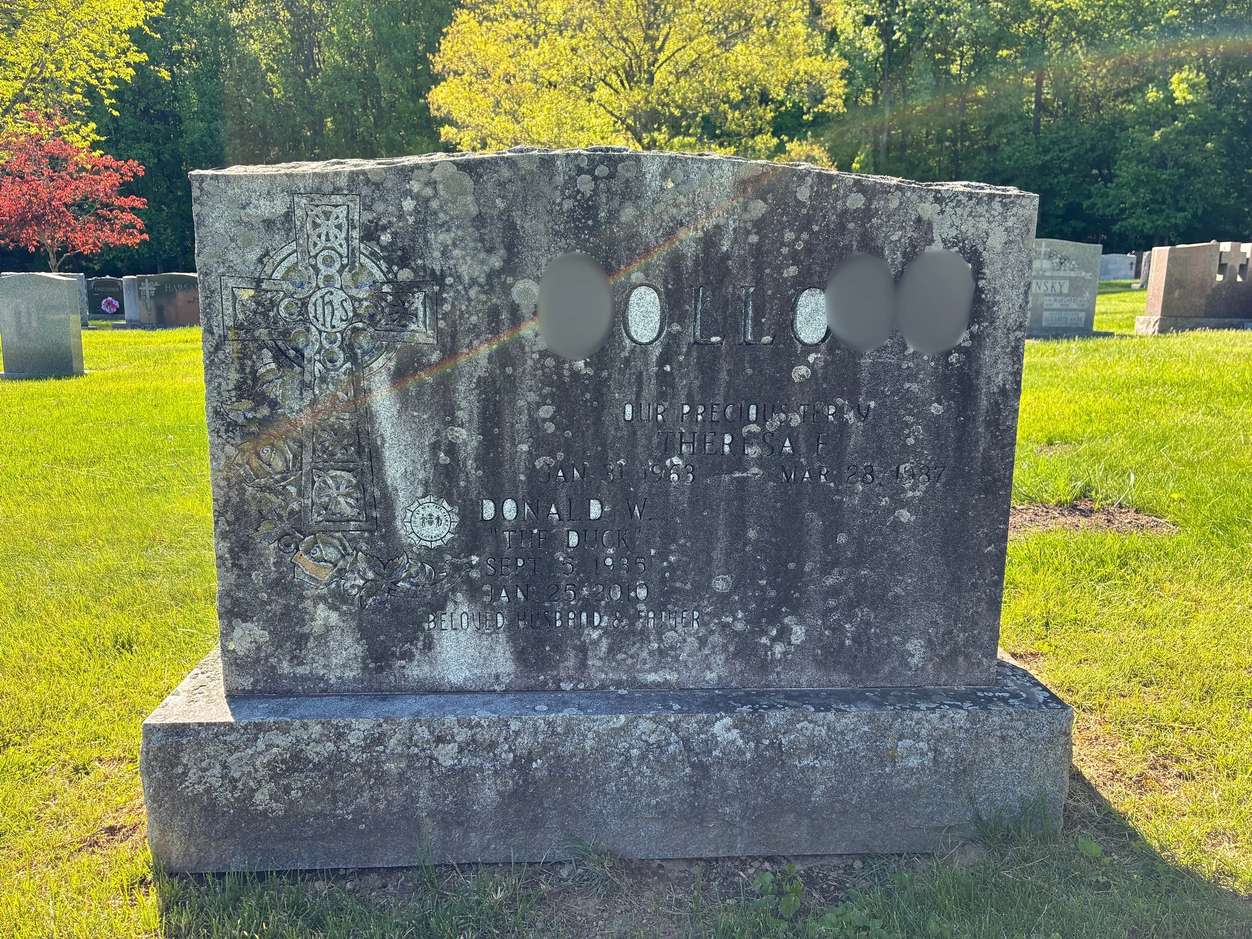 Gravestone in a cemetery with trees and grass, sunlight shining on it, and some text engraved on it.
