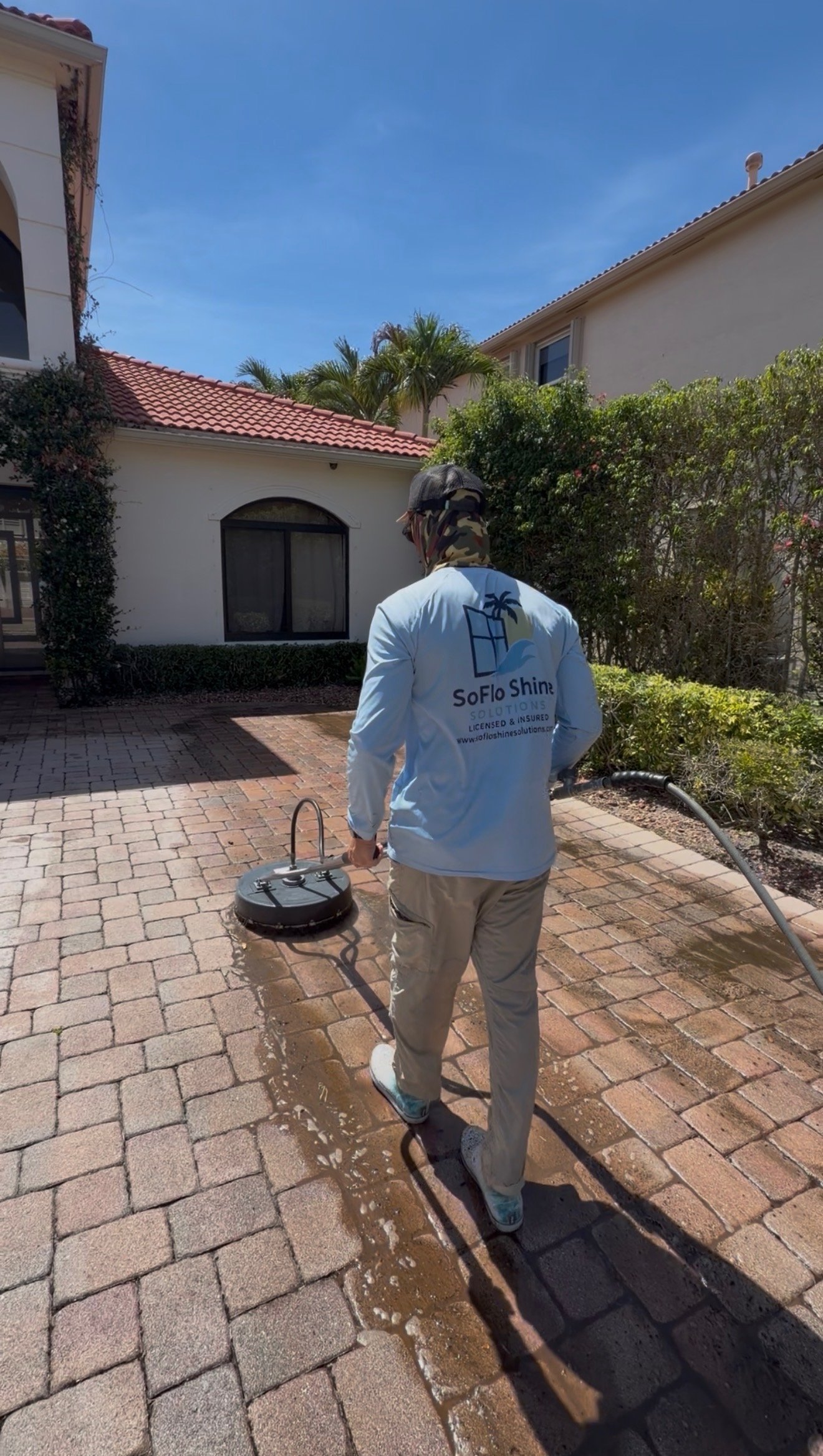 A person wearing a SoFlo Shine shirt cleaning a brick patio with a power washer on a sunny day, with residential houses and palm trees in the background.