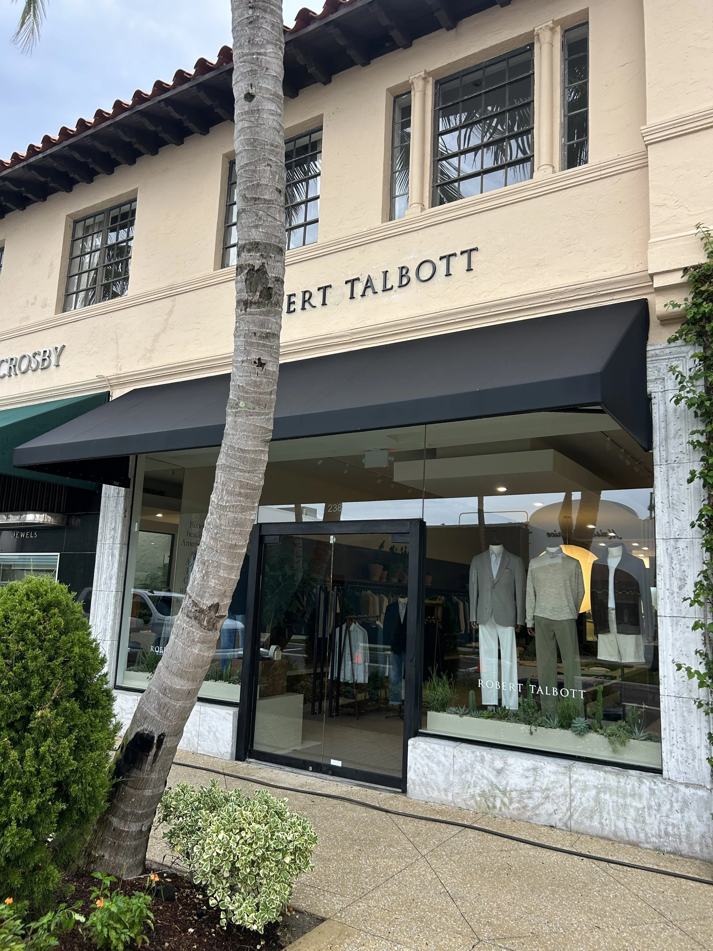 Storefront of Robert Talbott clothing boutique with large glass window displaying mannequins in suits and shirts, surrounded by greenery and a tree trunk in the foreground.