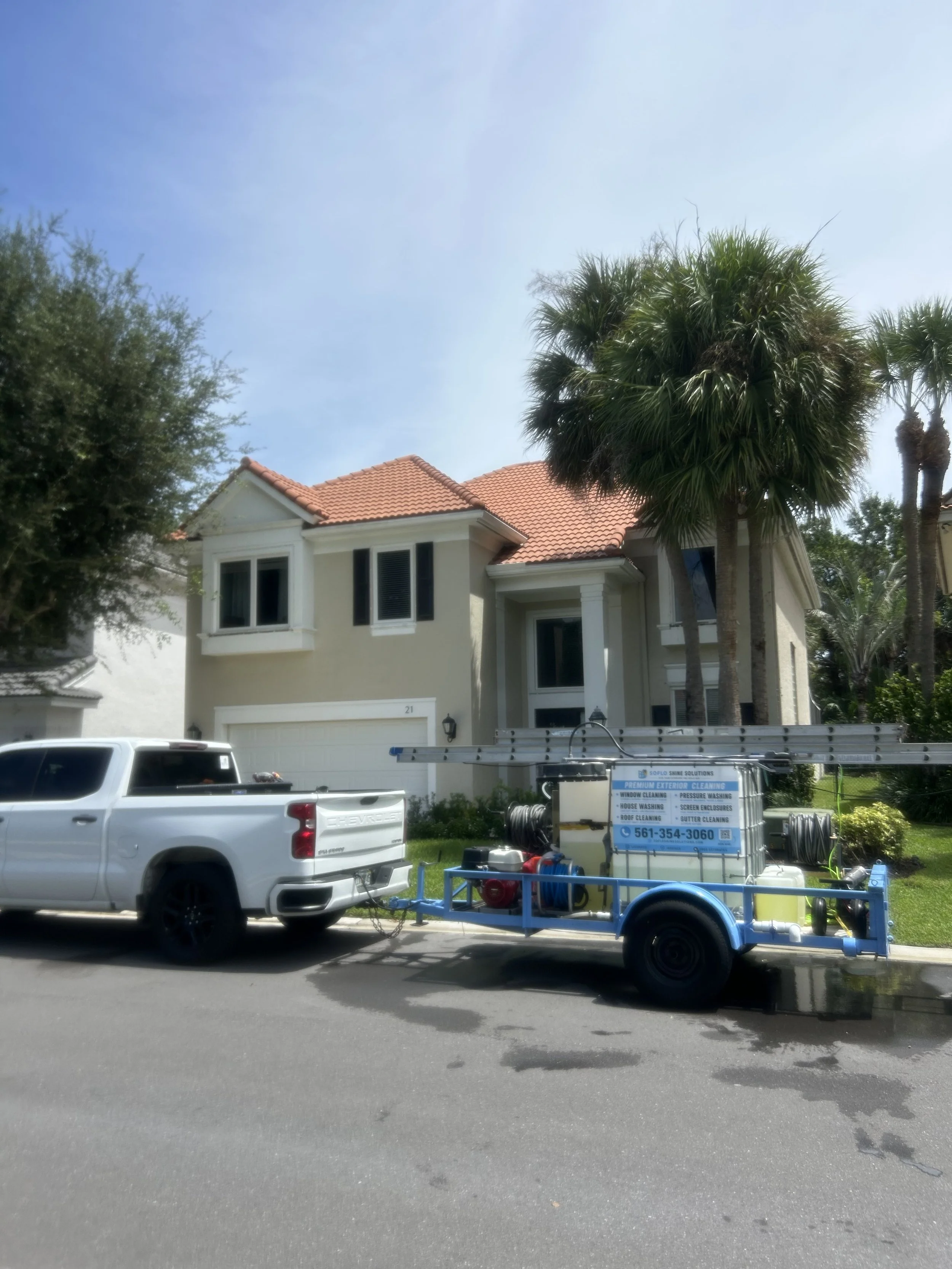 A white pickup truck parked on the street in front of a two-story house with a red-tiled roof, palm trees, and a lawn. There is a cleaning equipment trailer attached to the truck.