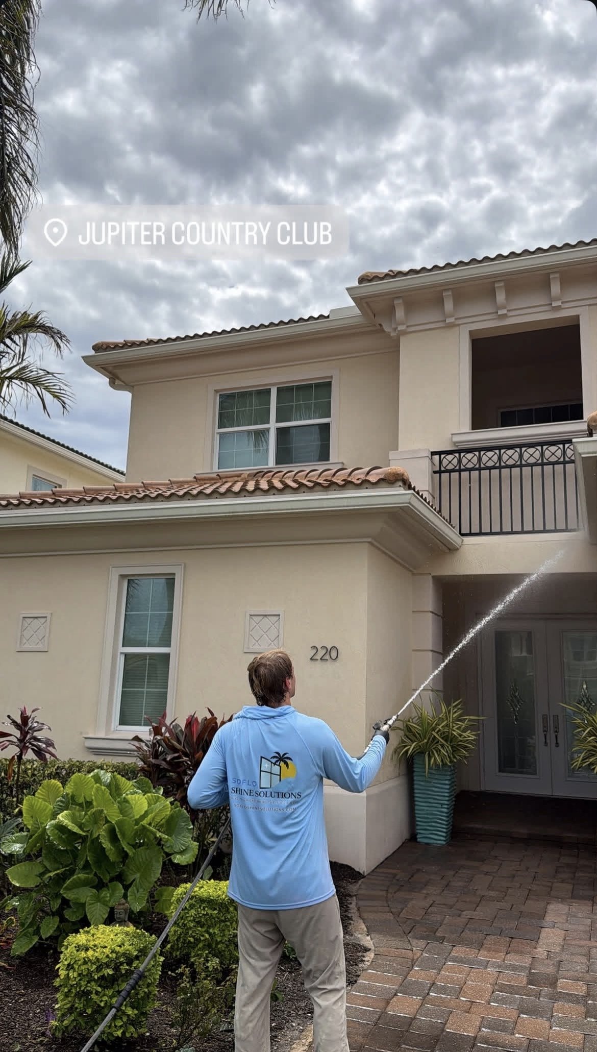 A man wearing a blue shirt with the logo 'SOFLO Shine Solutions' is watering plants in front of a yellow house with the number 220 on the wall. The house has beige walls, several windows, and a black metal balcony. The background shows a cloudy sky.