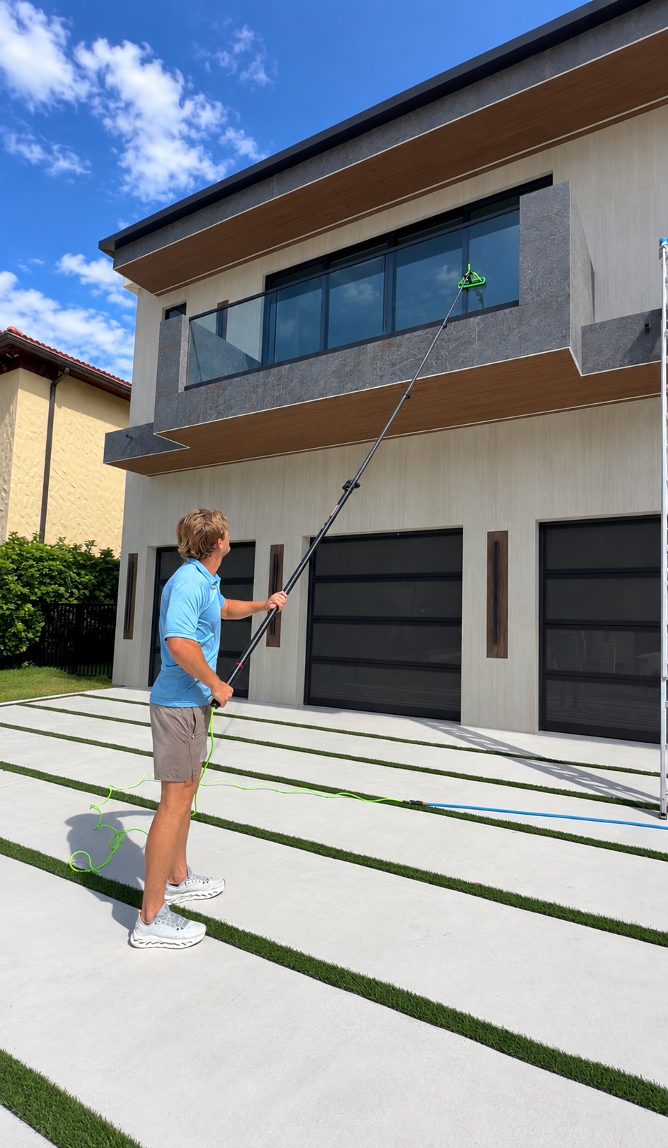 A man stands outside a modern house, holding a long pole with a green device at the end, reaching towards the balcony of the house under a blue sky with scattered clouds.