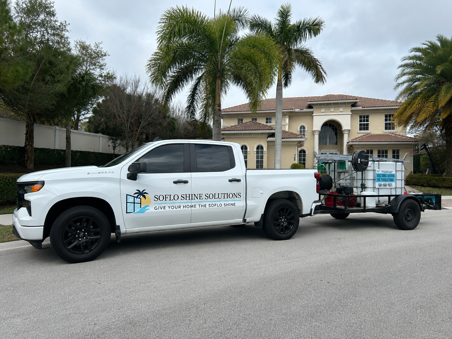 A white pickup truck with a logo and text that reads "SOFLO SHINE SOLUTIONS" and "GIVE YOUR HOME THE SOFLO SHINE" on the side. The truck is attached to a trailer carrying cleaning or pressure washing equipment, parked in front of a large beige house 