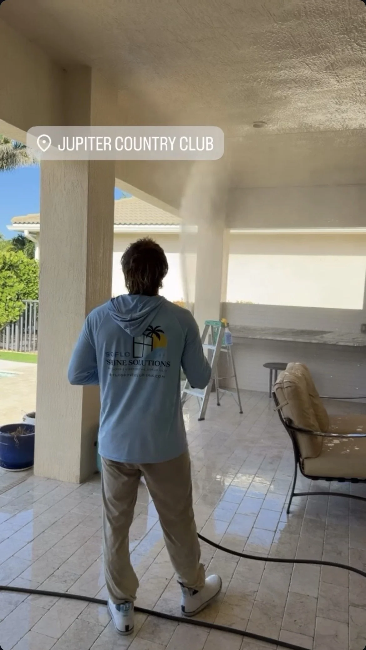 A person standing on a patio in front of a screened porch, using a high-pressure washer to clean the ceiling of the porch. The person is wearing a blue hoodie with a logo, beige pants, and white sneakers.