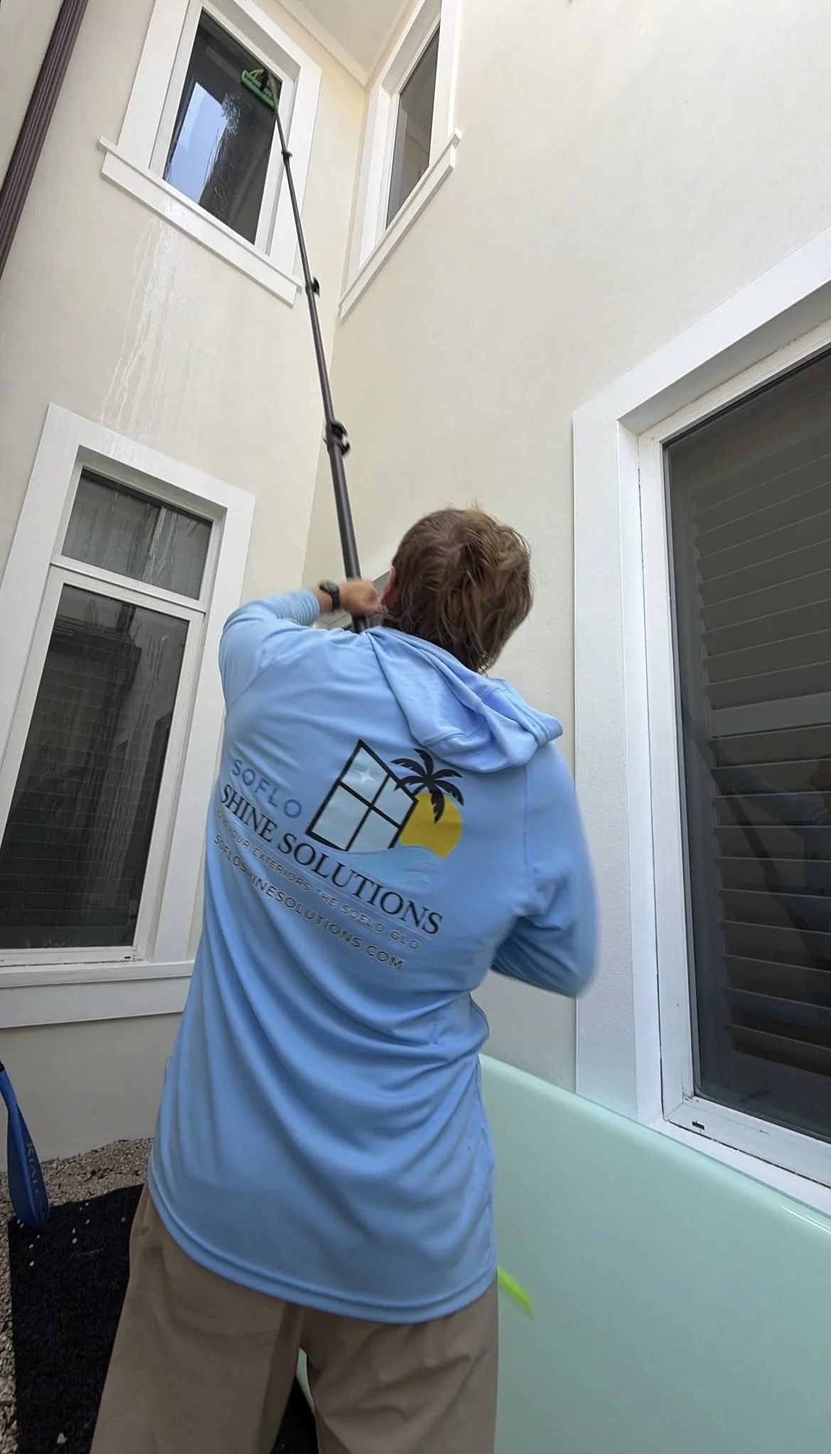 A person in a blue shirt using a water-fed pole to clean a second-story window on a beige house with white trim.