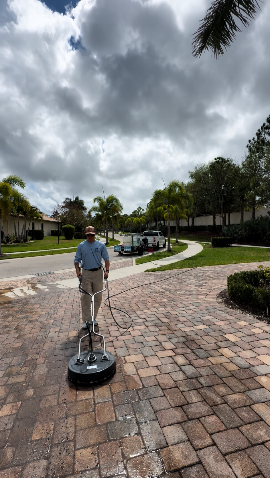 A man operating a floor cleaning machine on a paved driveway in a suburban neighborhood with palm trees, houses, and a cloudy sky overhead.