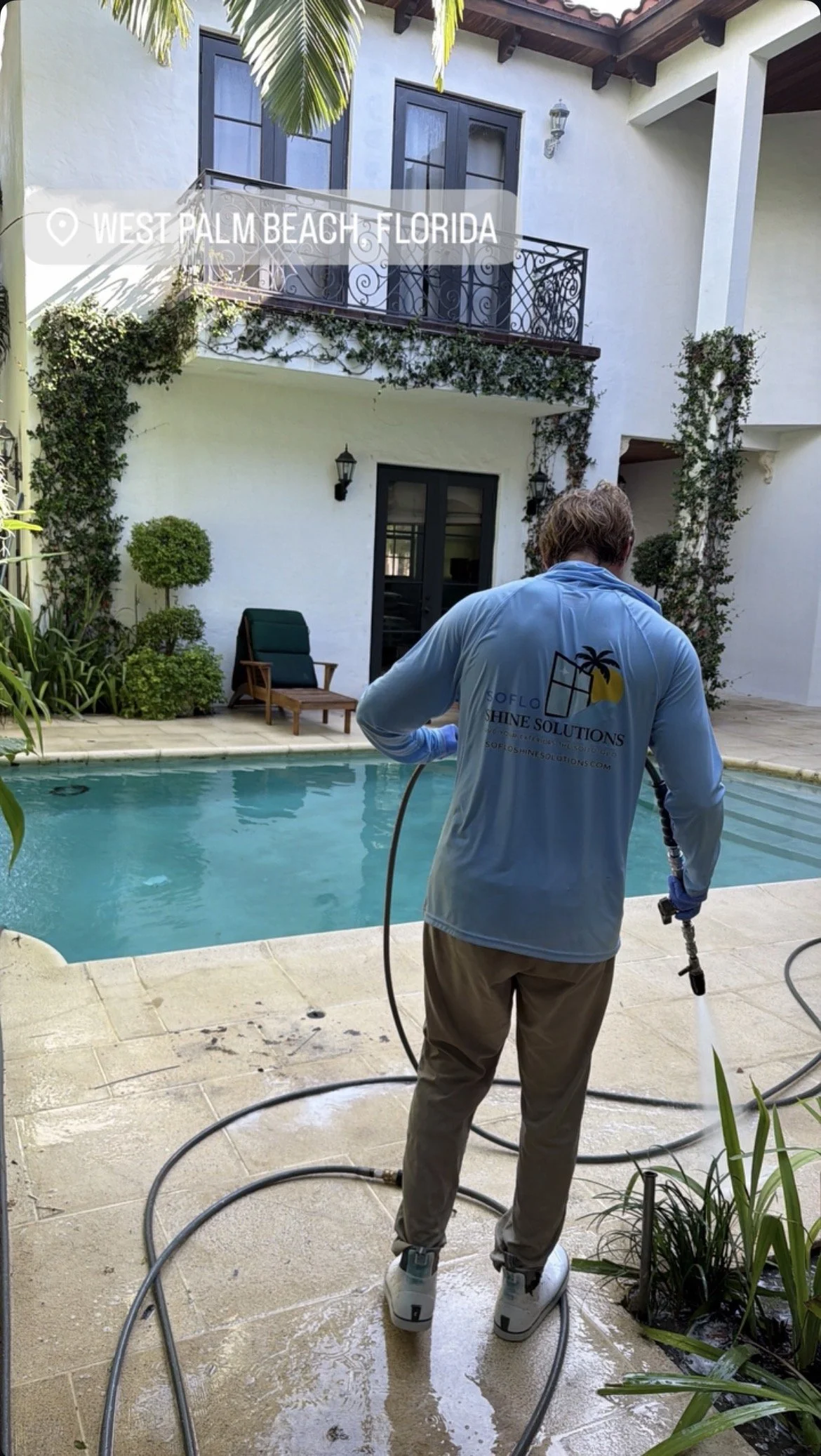 A man pressure washing a pool deck in the backyard of a house in West Palm Beach, Florida. The house has white stucco walls, black window frames, and a small balcony with iron railings. There are green plants and outdoor furniture around the pool are