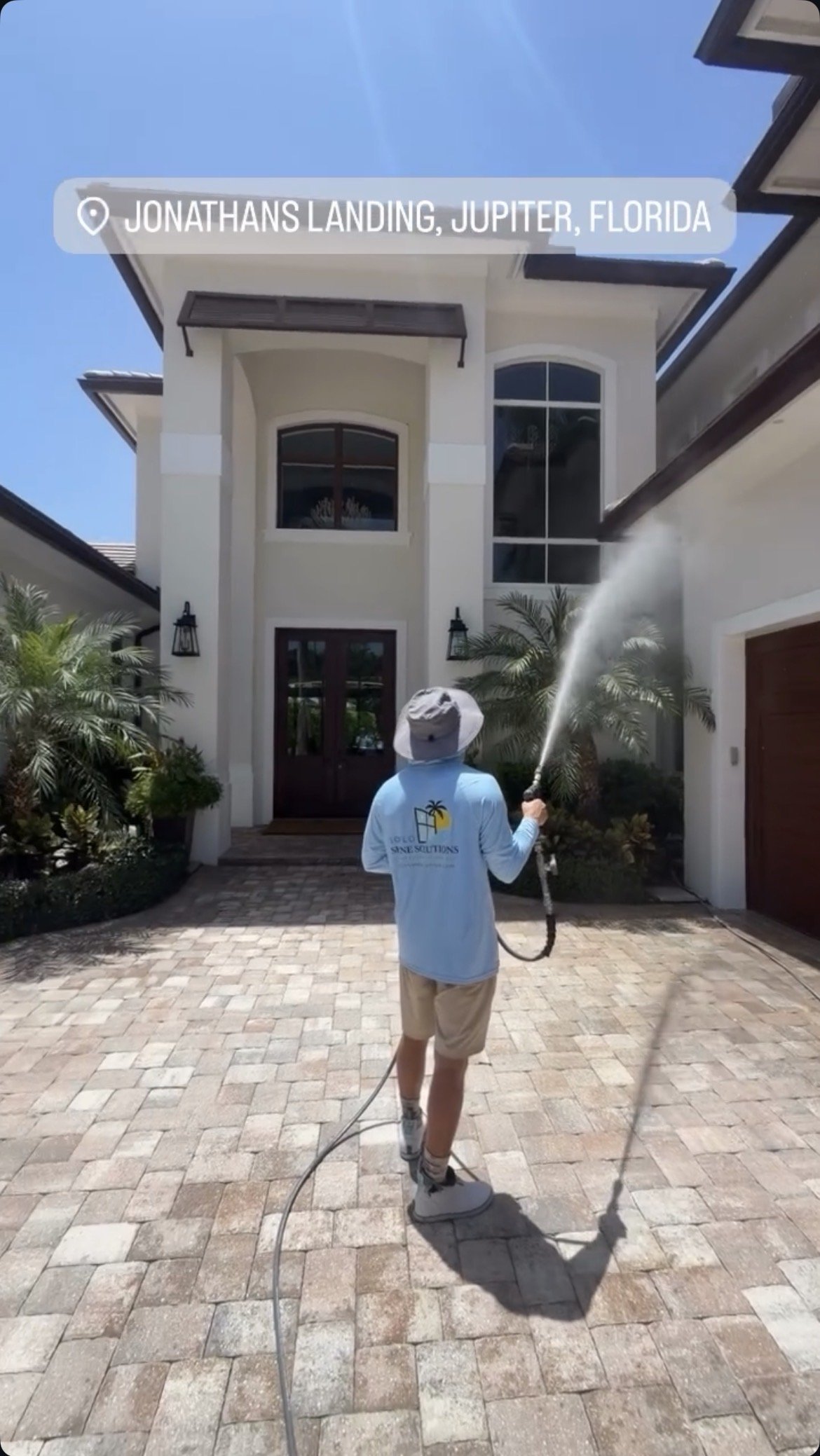 A person wearing a wide-brim hat and a long-sleeve shirt with a logo, holding a hose, watering or cleaning the front yard of a modern house with white exterior walls, large windows, palm trees, and a paved driveway in Jupiter, Florida.