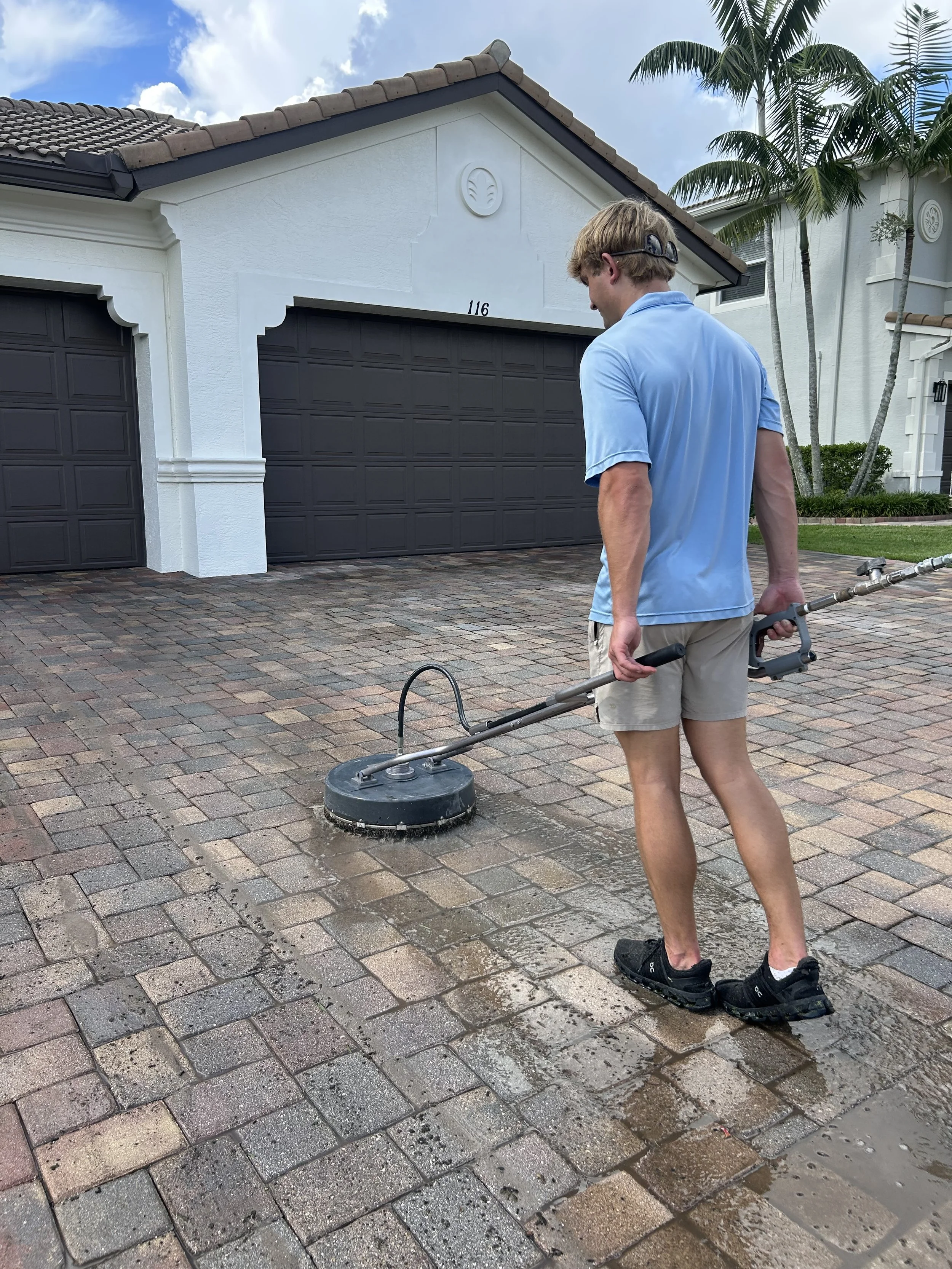 A young man in a light blue shirt and beige shorts is pressure washing a brick driveway. He is holding a pressure washer wand, cleaning the surface near a white house with a gray garage door. The scene is outdoors on a sunny day with a partly cloudy 