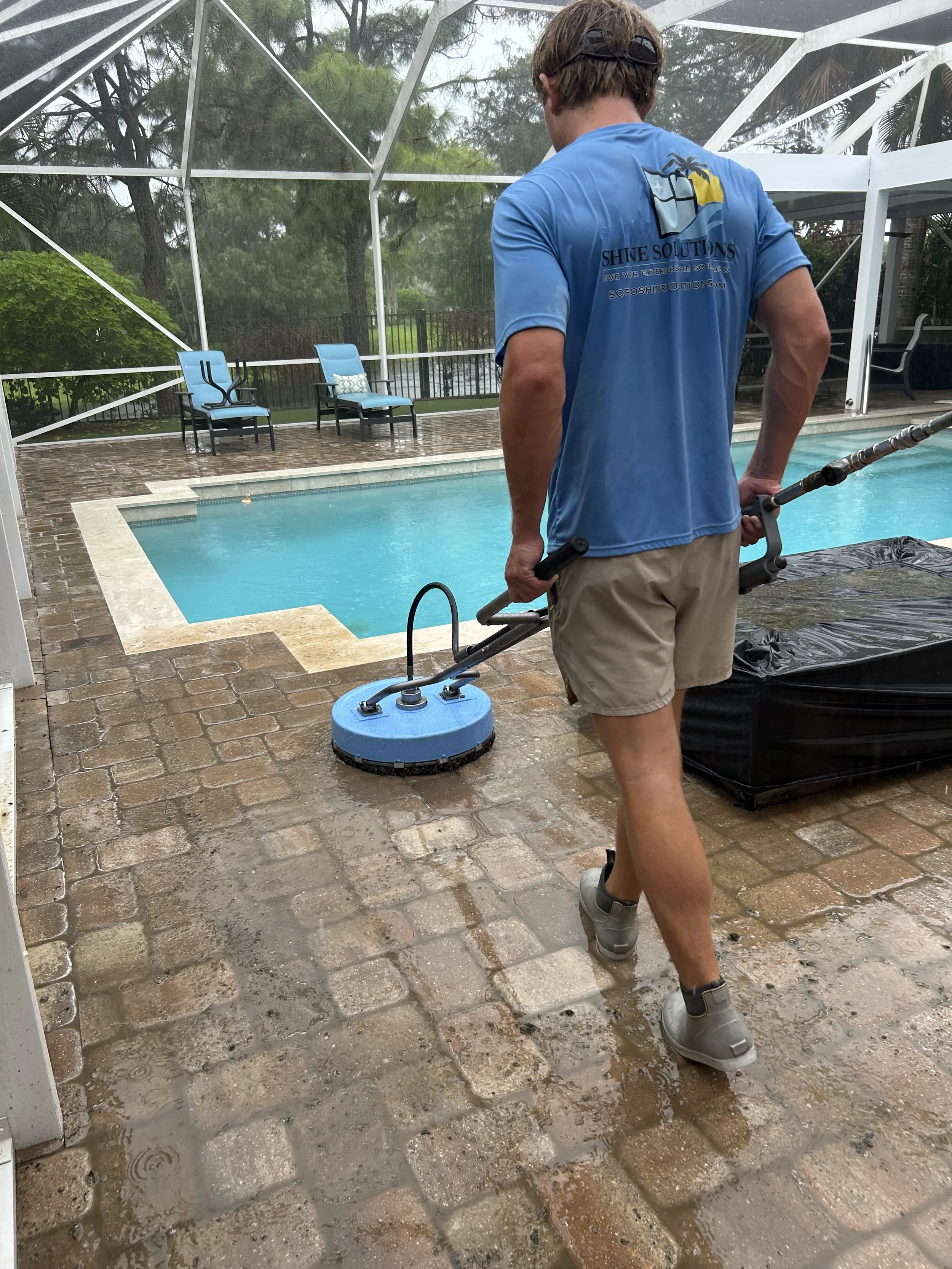 Man cleaning pool deck with a power washer on a wet brick patio, patio furniture in background, enclosed pool area with trees outside.