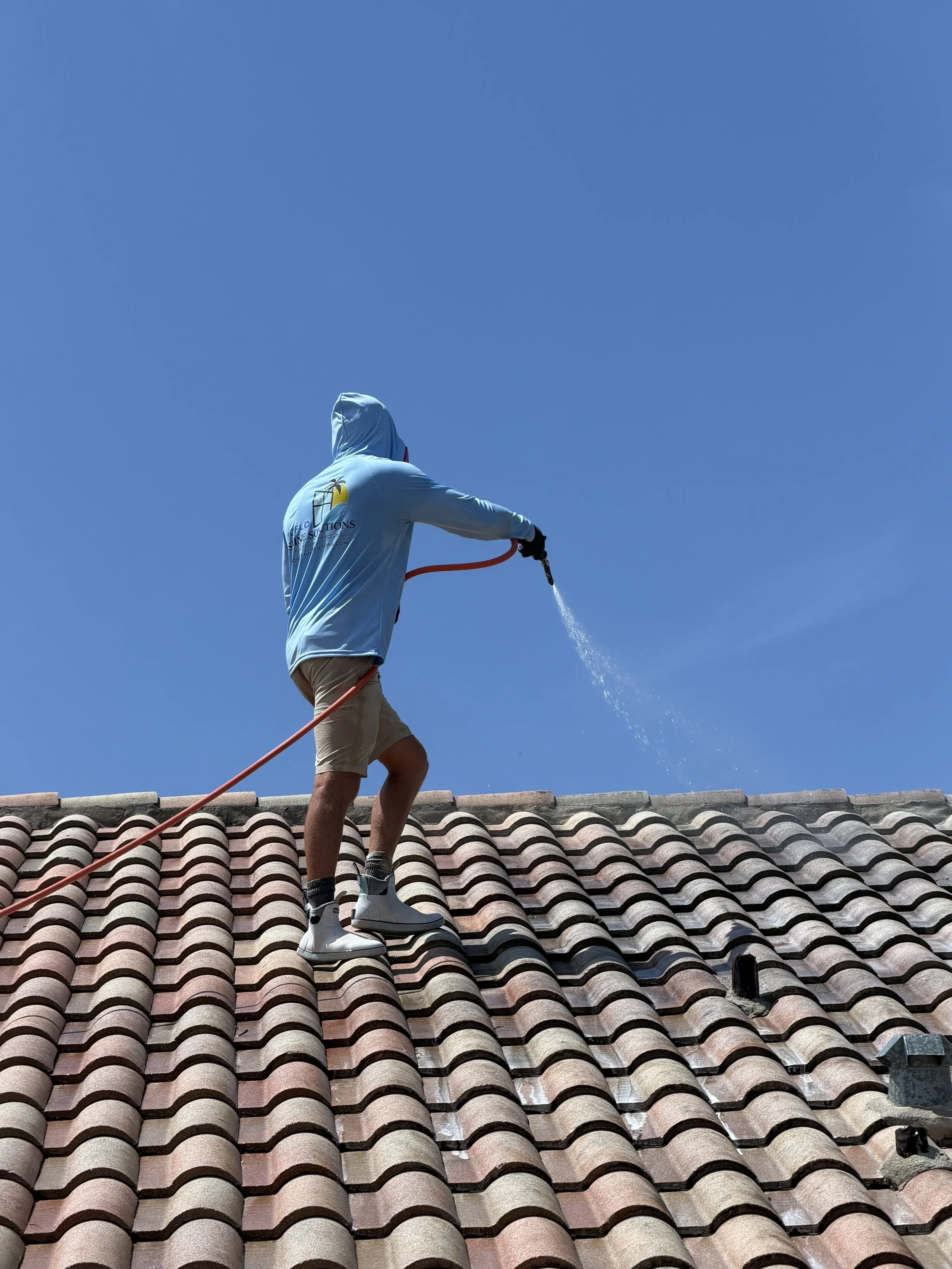 A person wearing a light blue hoodie with a logo, khaki shorts, and white sneakers is standing on a tiled roof, spraying water with a hose against a clear blue sky.