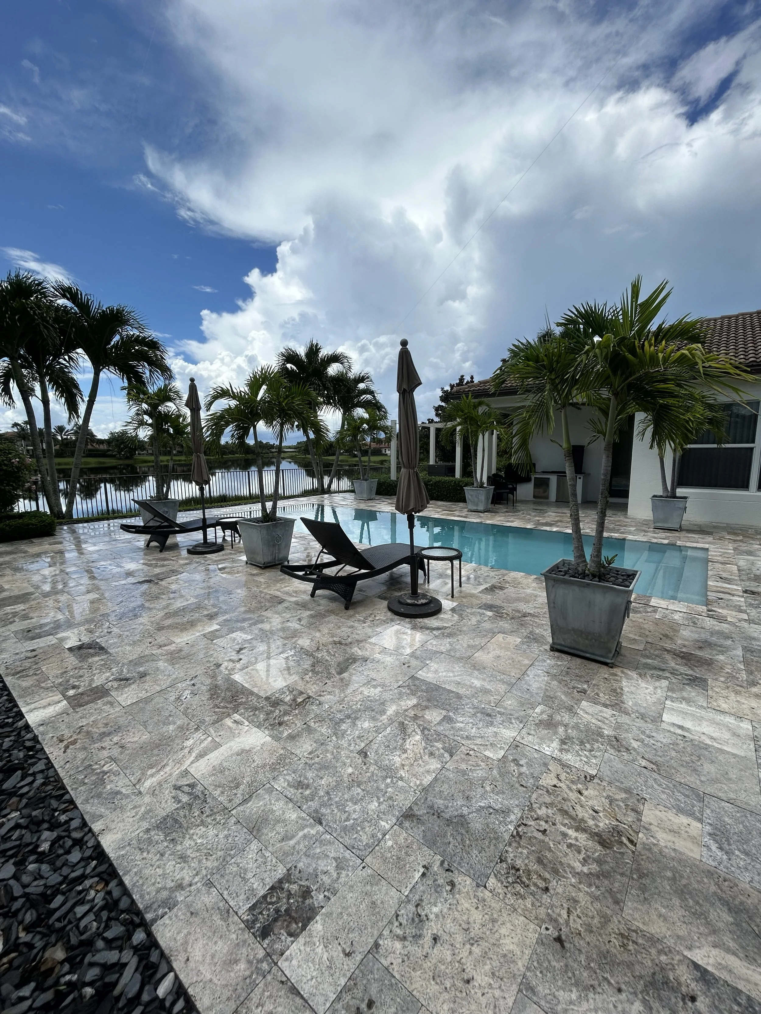 View of a backyard patio with a swimming pool, lounge chairs, closed umbrellas, potted palm trees, a fence, and a house with a tile roof, under a partly cloudy sky.