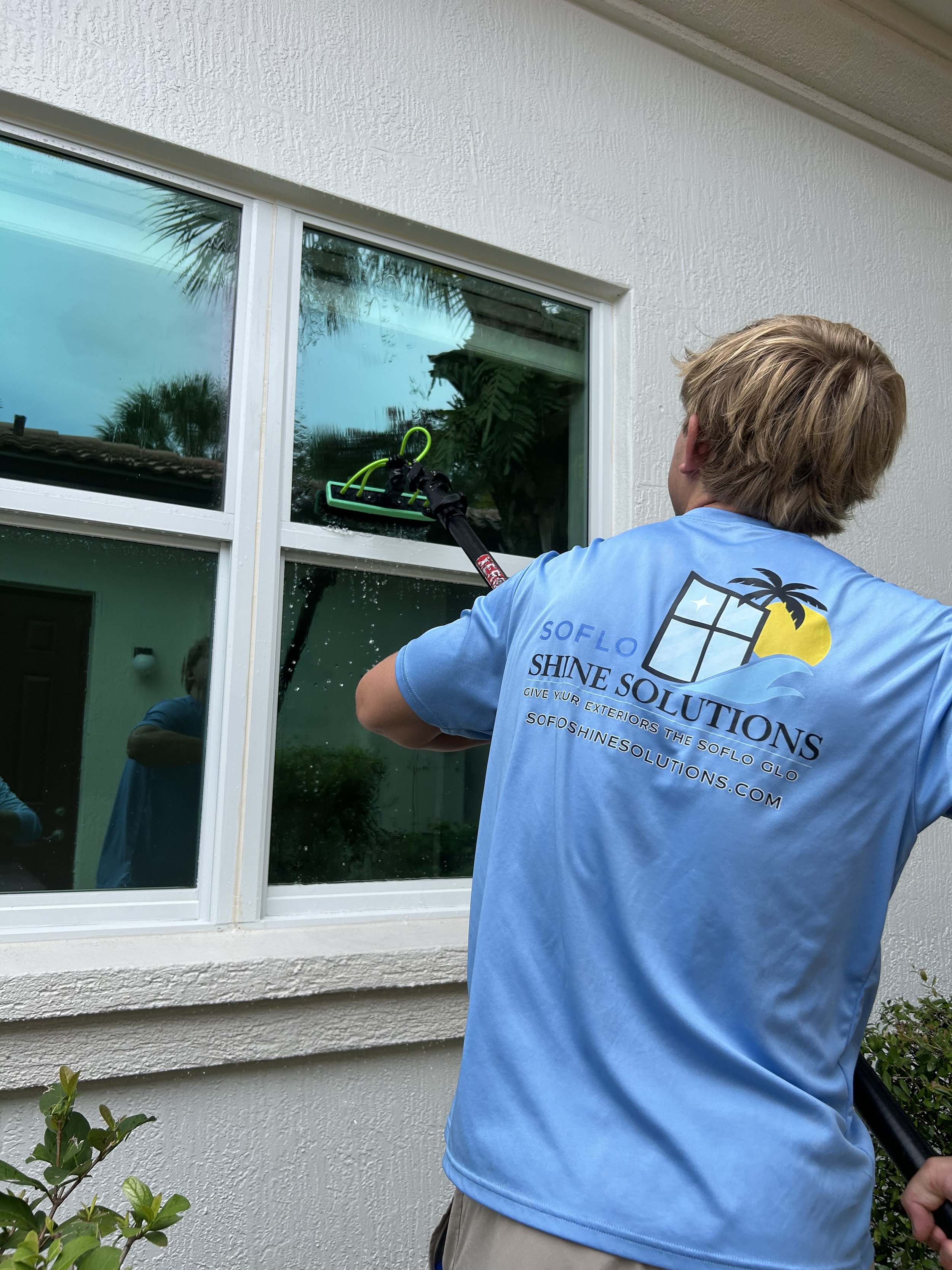 A person cleaning a window with a power washer on a white wall with a window and some greenery in the foreground.