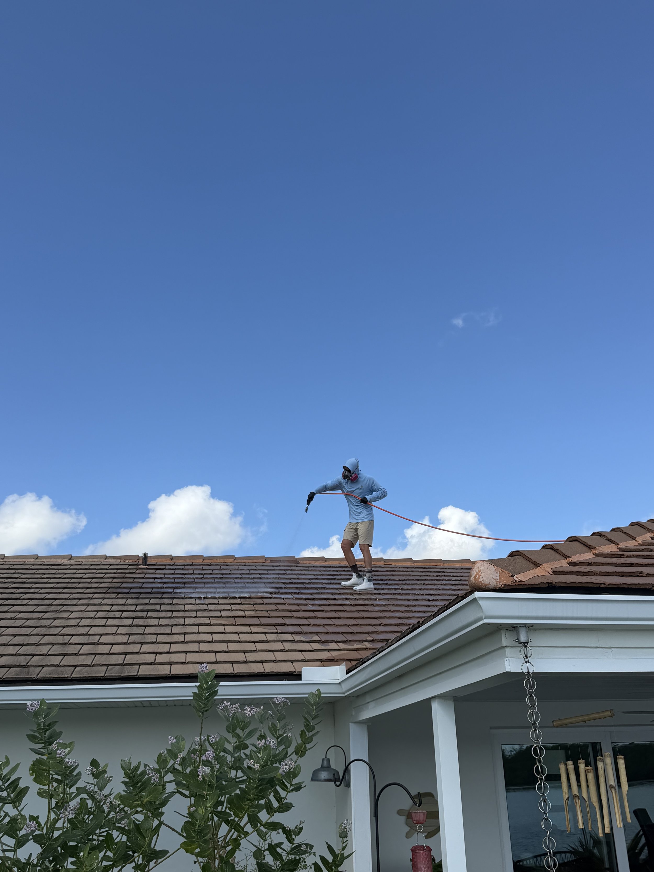 A person wearing a long sleeve shirt, shorts, a cap, and a mask is standing on a rooftop, holding a long tool, and working on the roof tiles. The sky is blue with some clouds, and part of a house's patio area with plants, lights, and a window is visi
