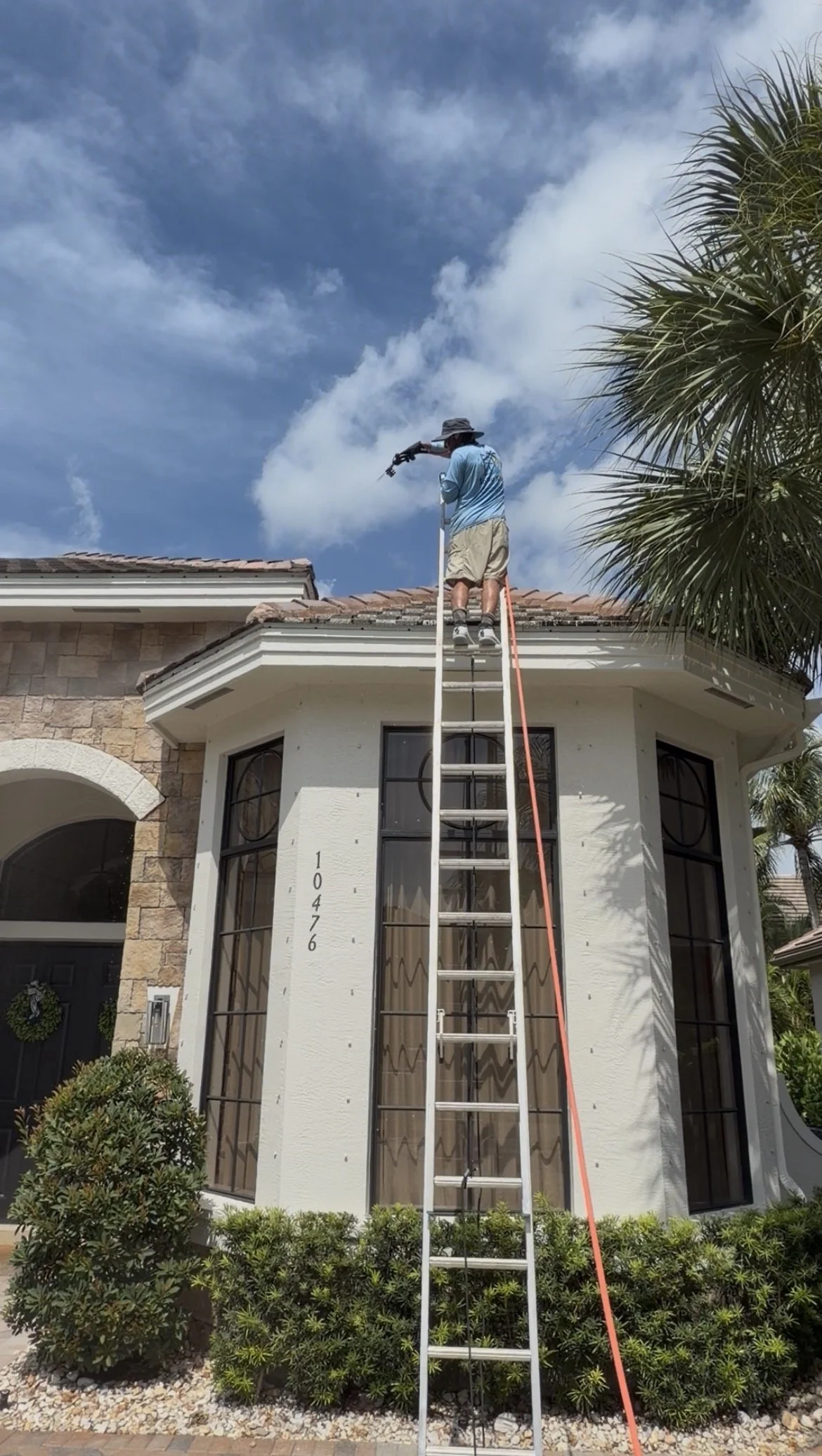 A man standing on a ladder cleaning or inspecting the roof of a house on a sunny day with a partly cloudy sky. The house has large windows and a front garden with bushes and decorative rocks.