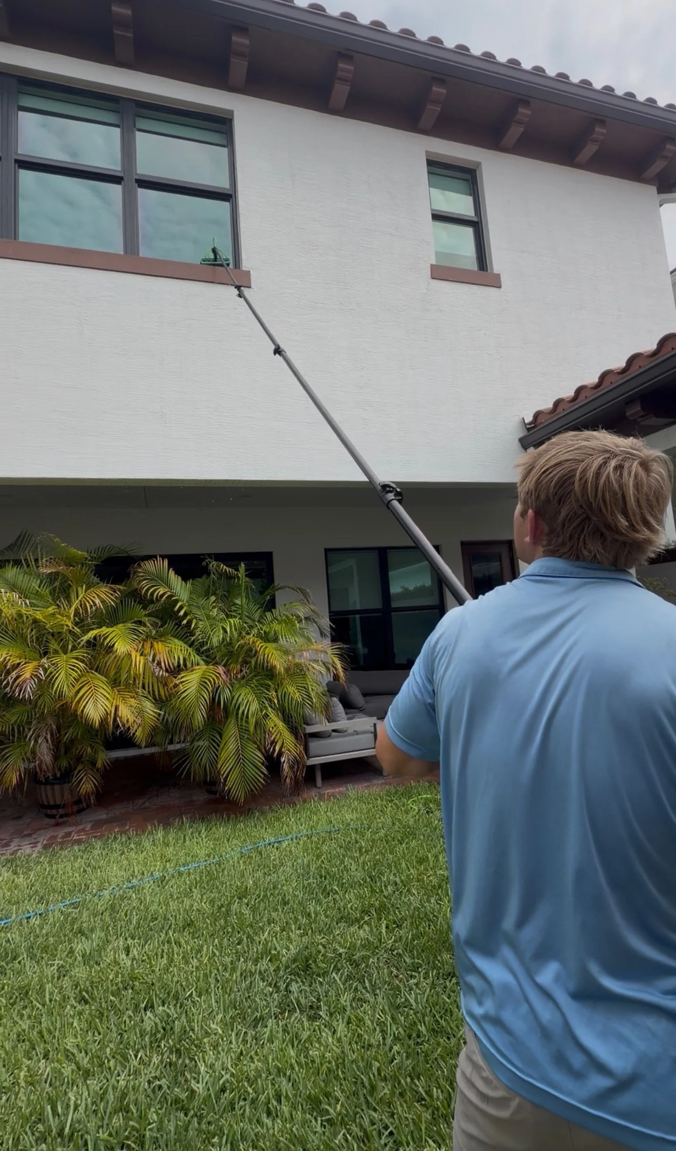 A man in a blue shirt is holding a long pole and cleaning a large window on the upper floor of a house. The house has a white exterior with brown trim and a tiled roof. There are plants and outdoor furniture in the yard, and the sky is partly cloudy.