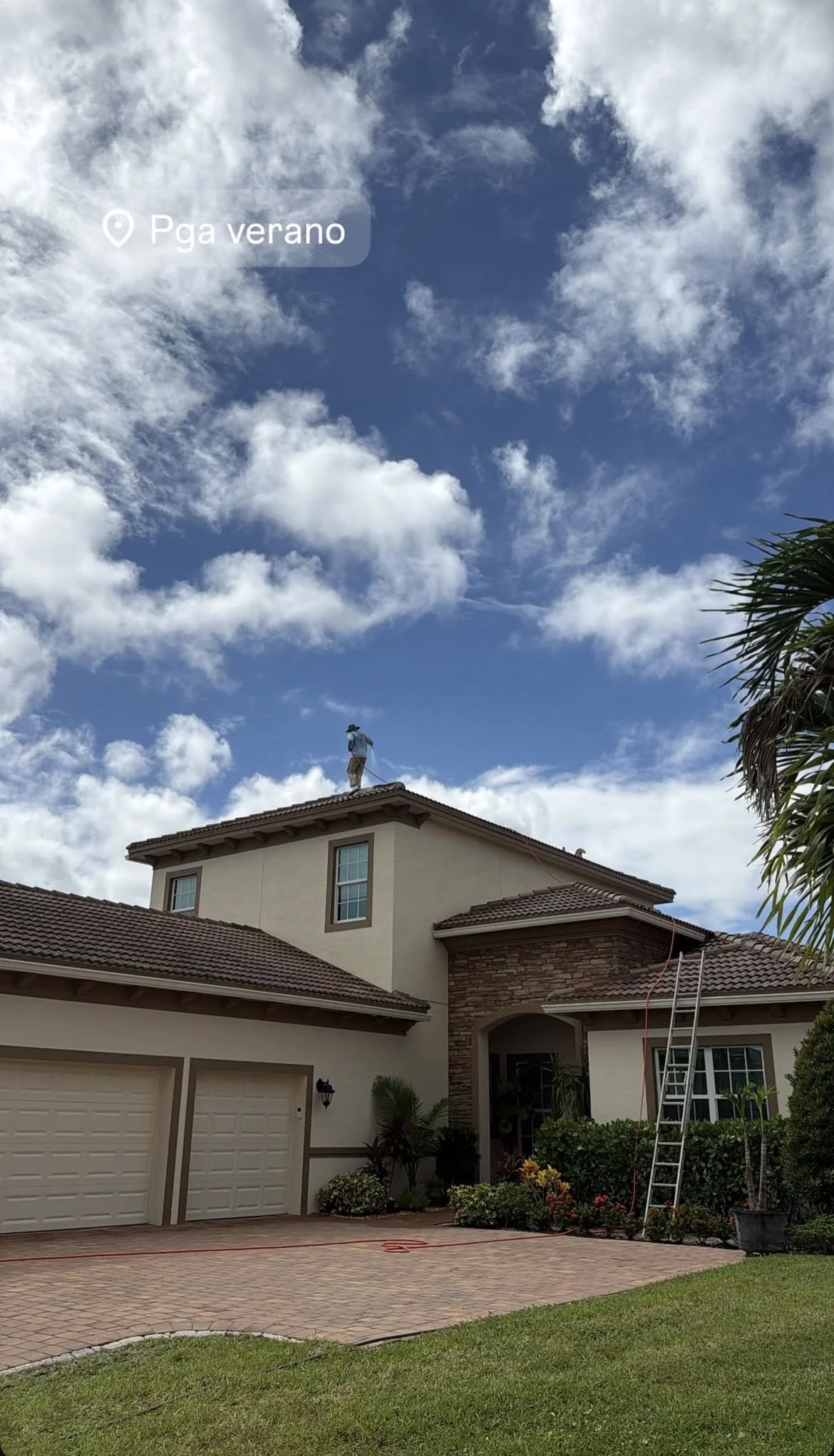 A person standing on the roof of a two-story house with a tiled roof, blue sky with clouds, and a palm tree on the right side.