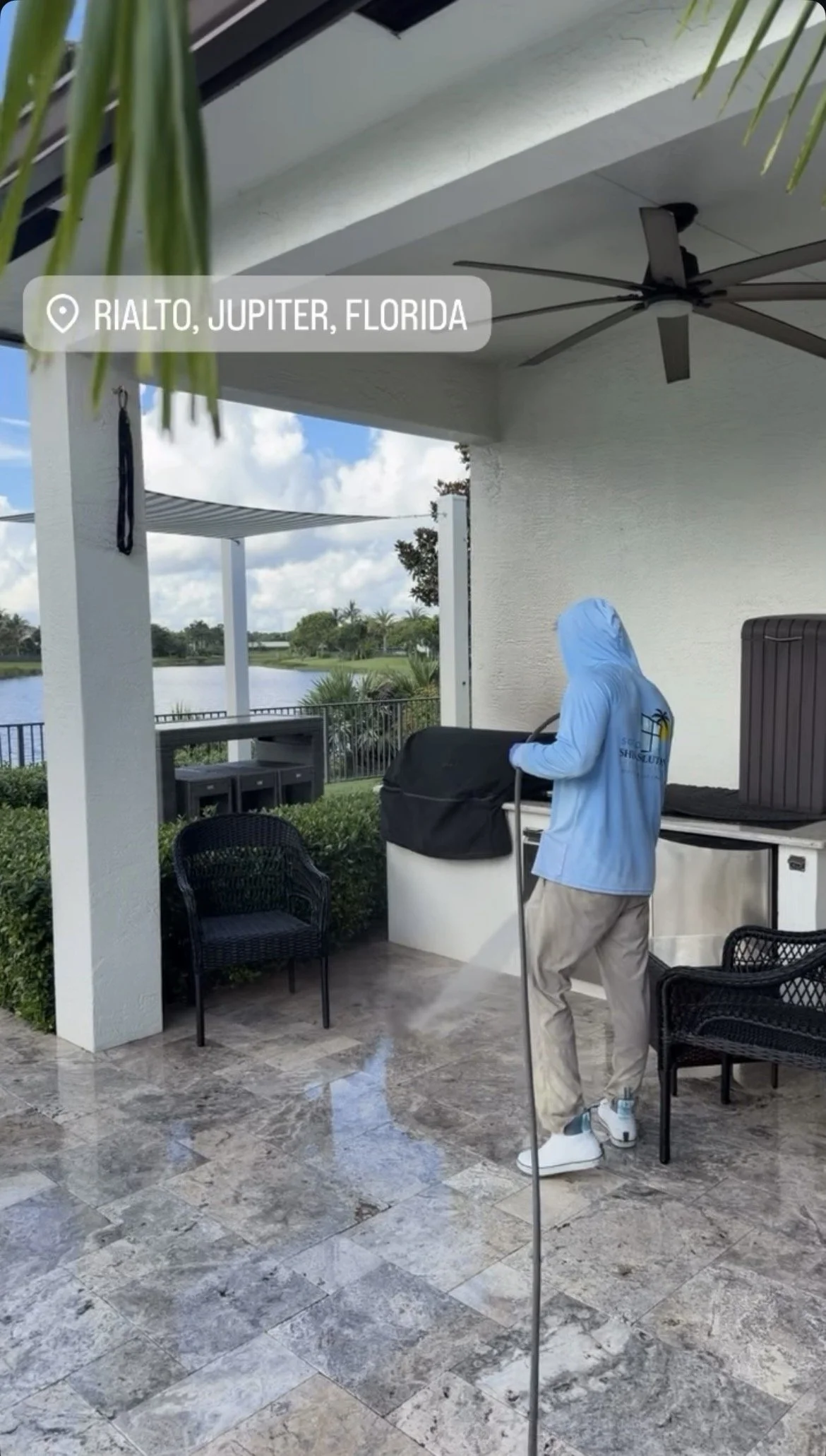A person wearing a blue hoodie, beige pants, and sneakers standing outside on a patio with a mop, near a small white refrigerator and black chairs. In the background, there is a lake, trees, and a partly cloudy sky. The caption indicates the location