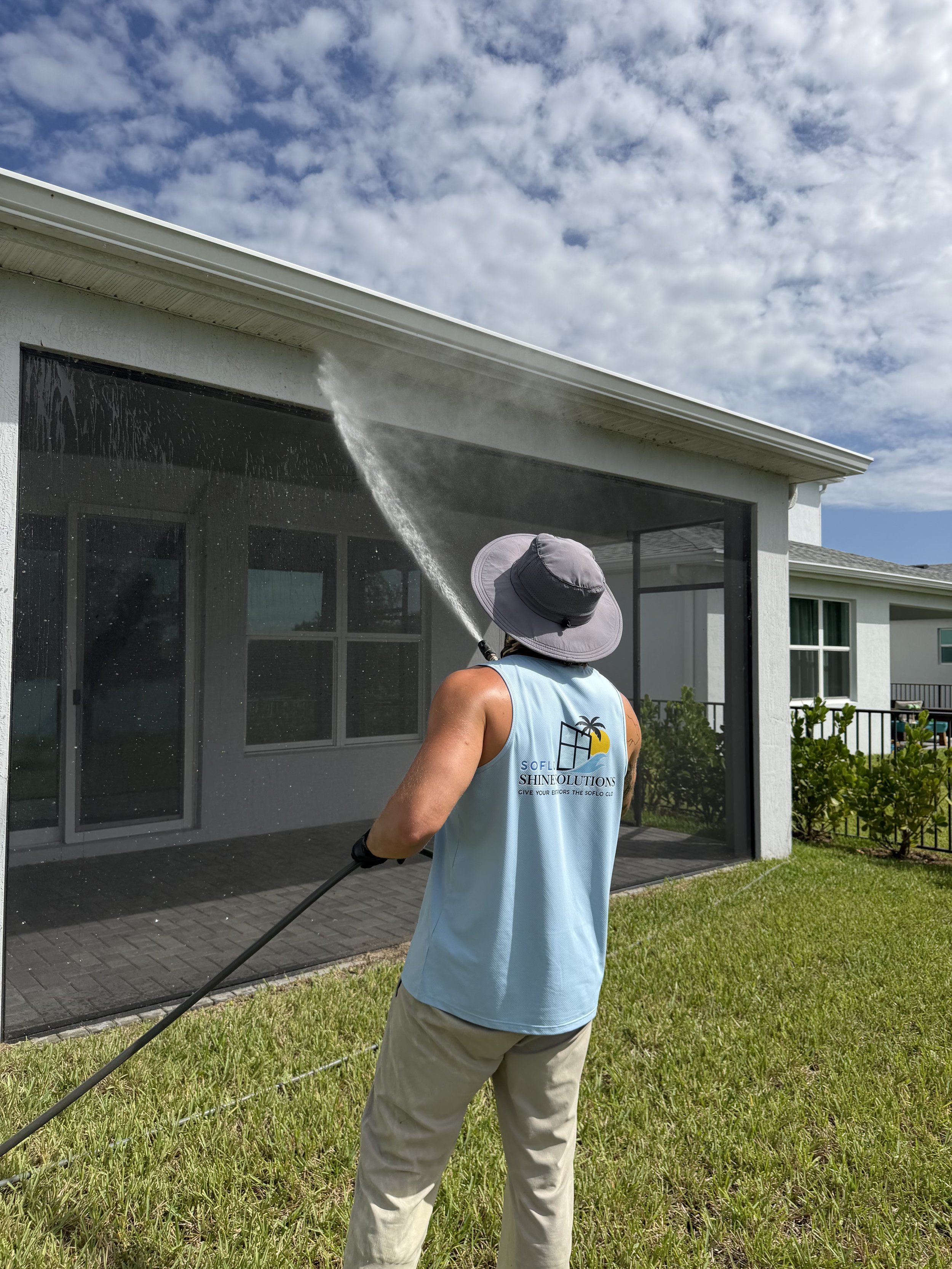 A person wearing a hat and a sleeveless shirt is using a pressure washer to clean the exterior of a screened-in porch or patio. The house has a white wall and a gray roof, and the background shows a partly cloudy sky.