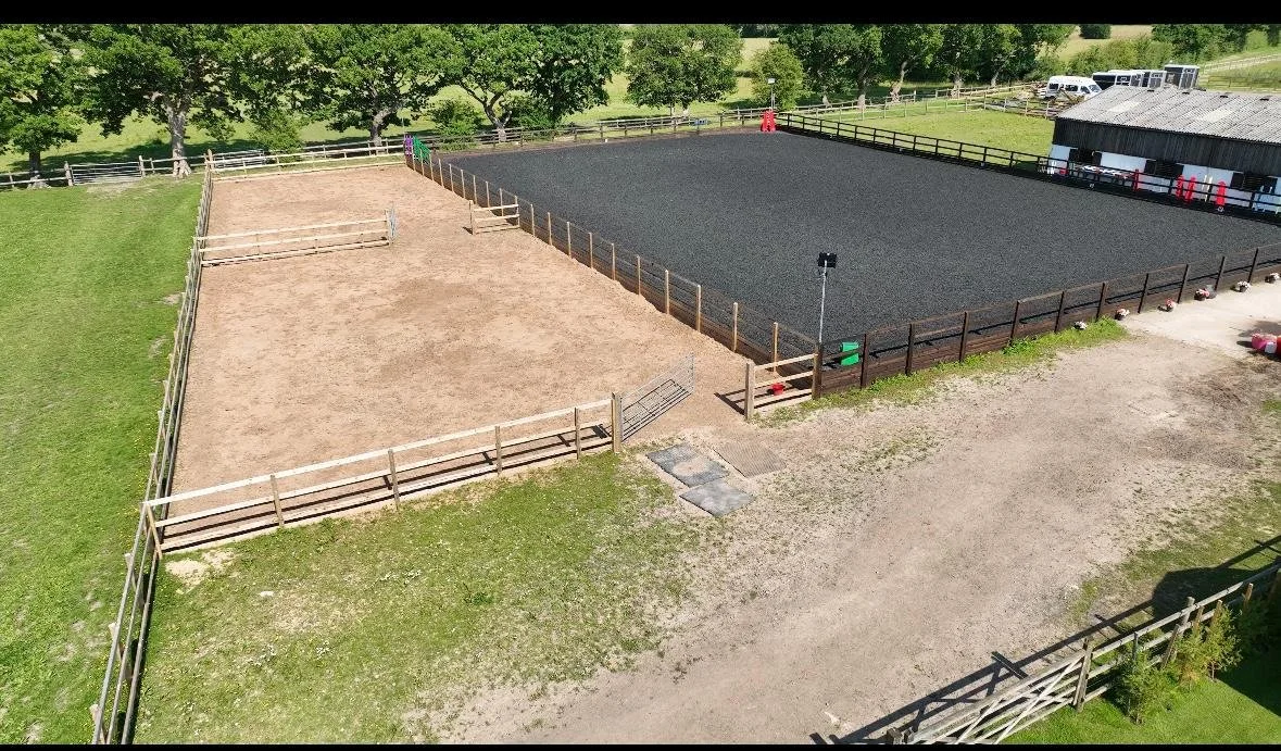 An outdoor equestrian riding arena with two sections, one with sand and the other with black footing, surrounded by wooden fences, with trees in the background and a barn to the right.