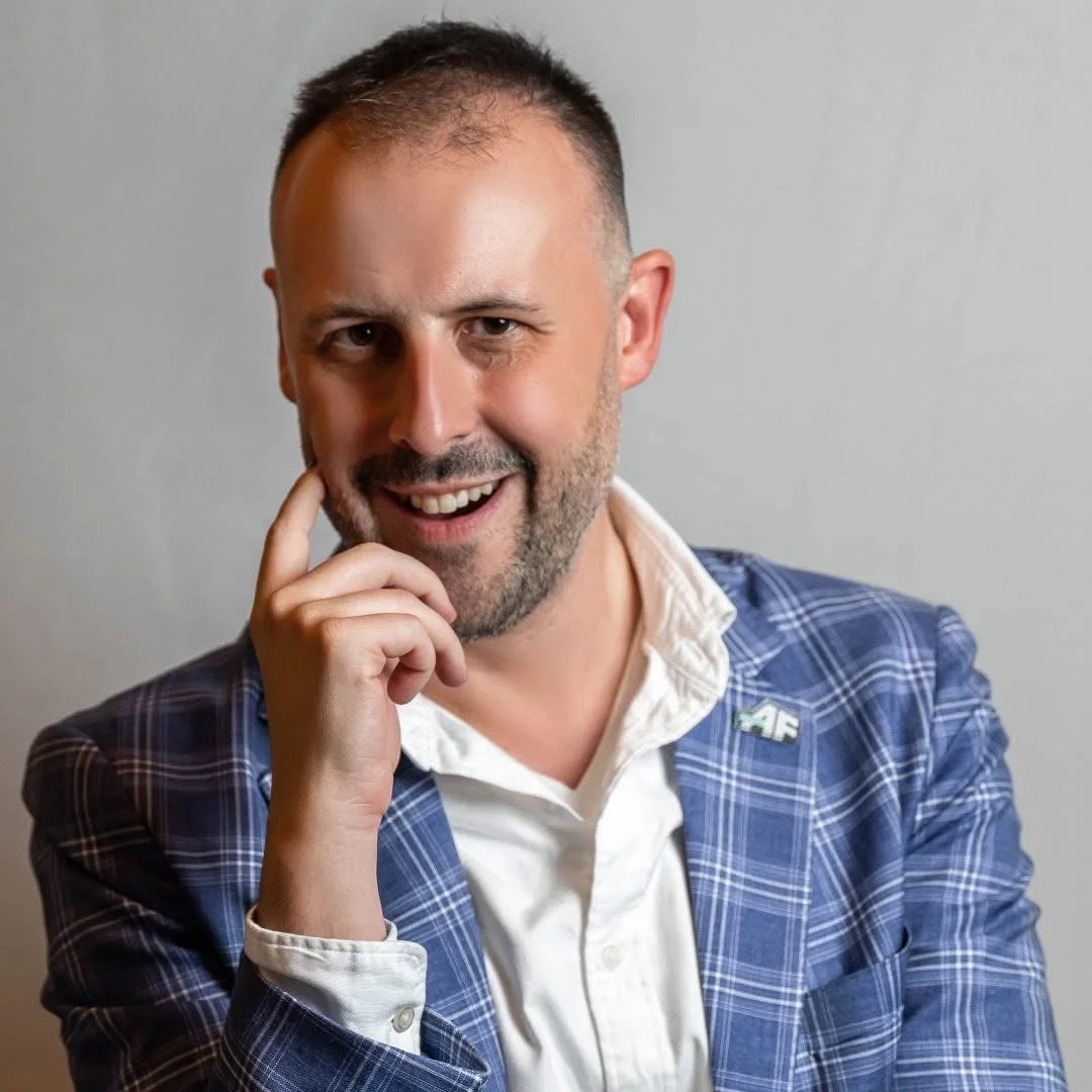 A smiling man with short dark hair, a beard, wearing a blue plaid suit jacket over a white shirt, with his hand resting on his chin, against a neutral background.