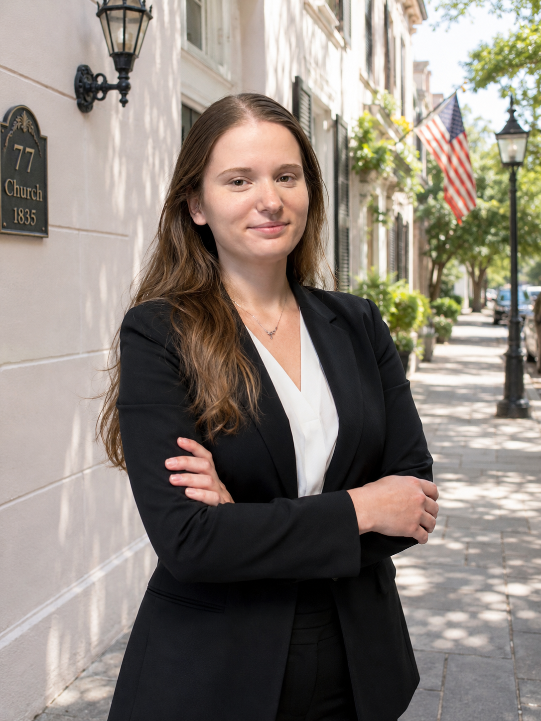 A woman with long brown hair, wearing a black blazer and white top, standing with arms crossed on a city sidewalk with American flags and greenery in the background.
