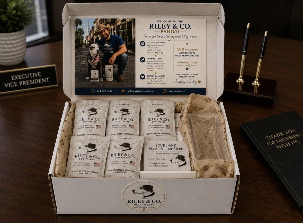 Open Riley & Co. dog treats gift box with five packets of peanut butter and blueberry flavored treats, a clear plastic bag, a promotional card, and a blank label for a bank. The box features a photograph of a man with a dog, and the interior includes informational text about the brand and their mission.