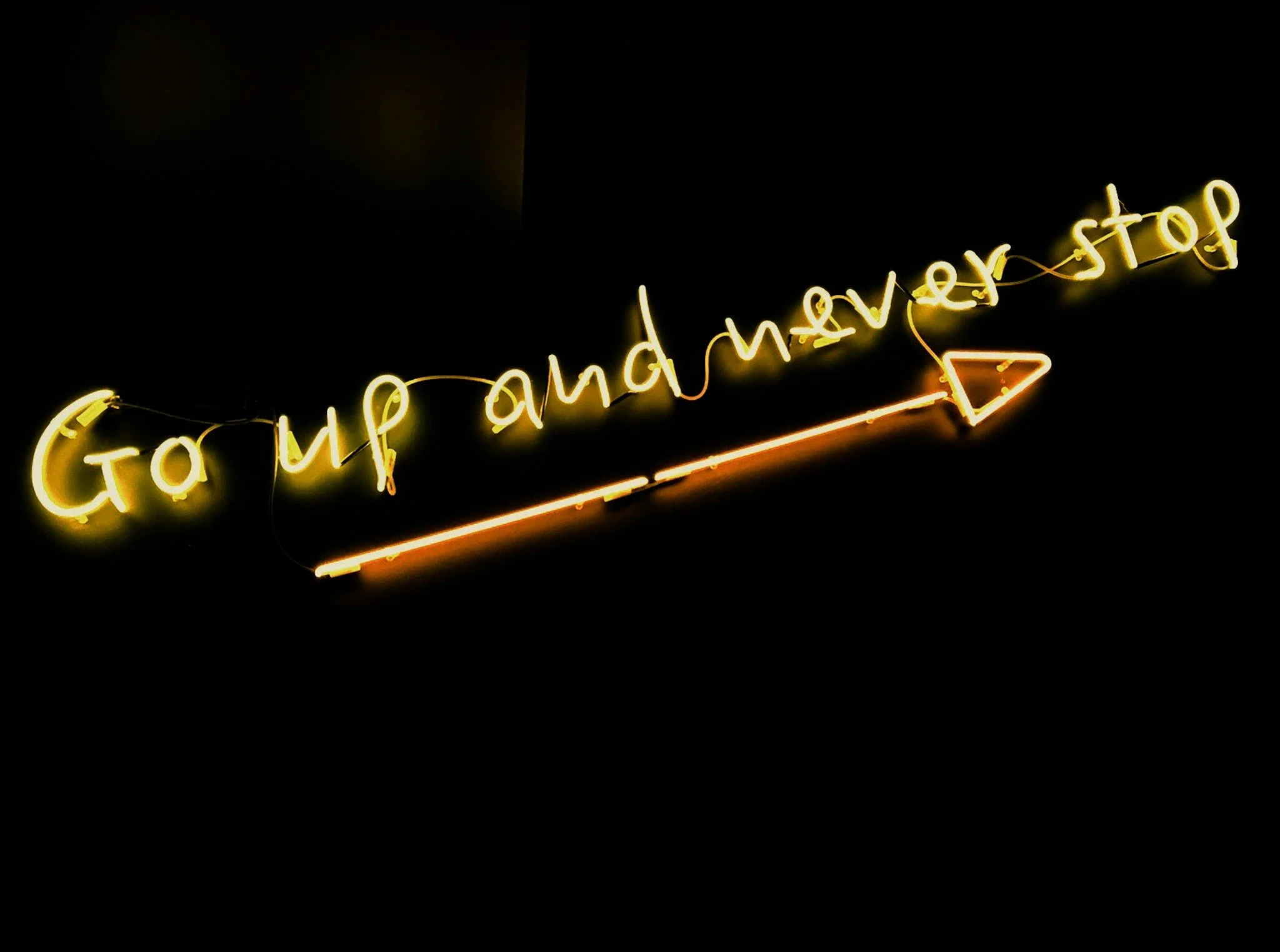 Neon sign with yellow text saying 'Go up and never stop' and a red arrow pointing to the right.