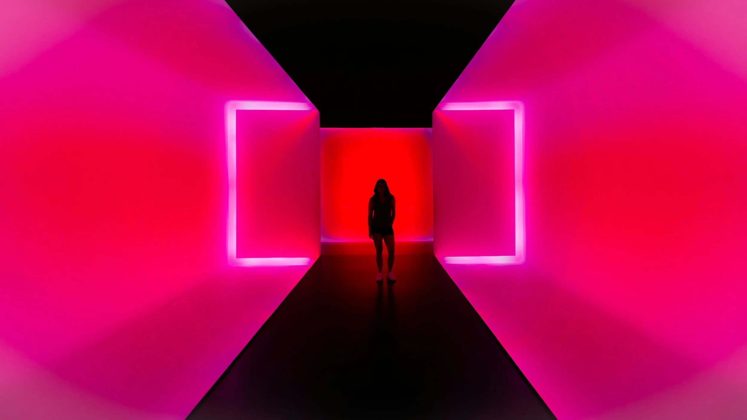 A person walking in a dimly lit corridor illuminated by vibrant pink and red neon lights, creating a futuristic atmosphere.
