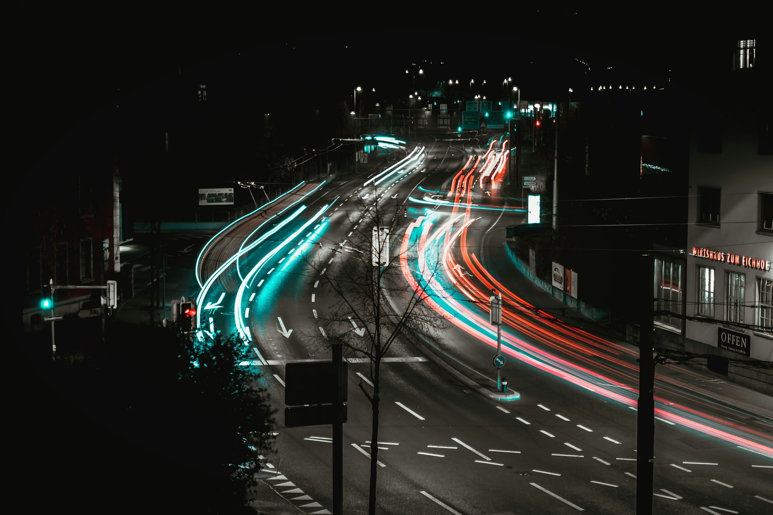 A long-exposure photo of a city street at night showing streaks of light from moving vehicles, with traffic lights and street signs visible.