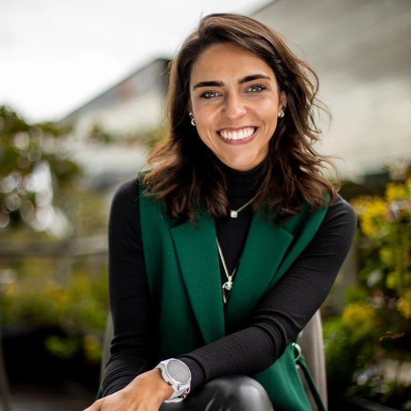 Smiling woman with brown hair wearing a black turtleneck and green vest, sitting outdoors with a blurred background of greenery and buildings.