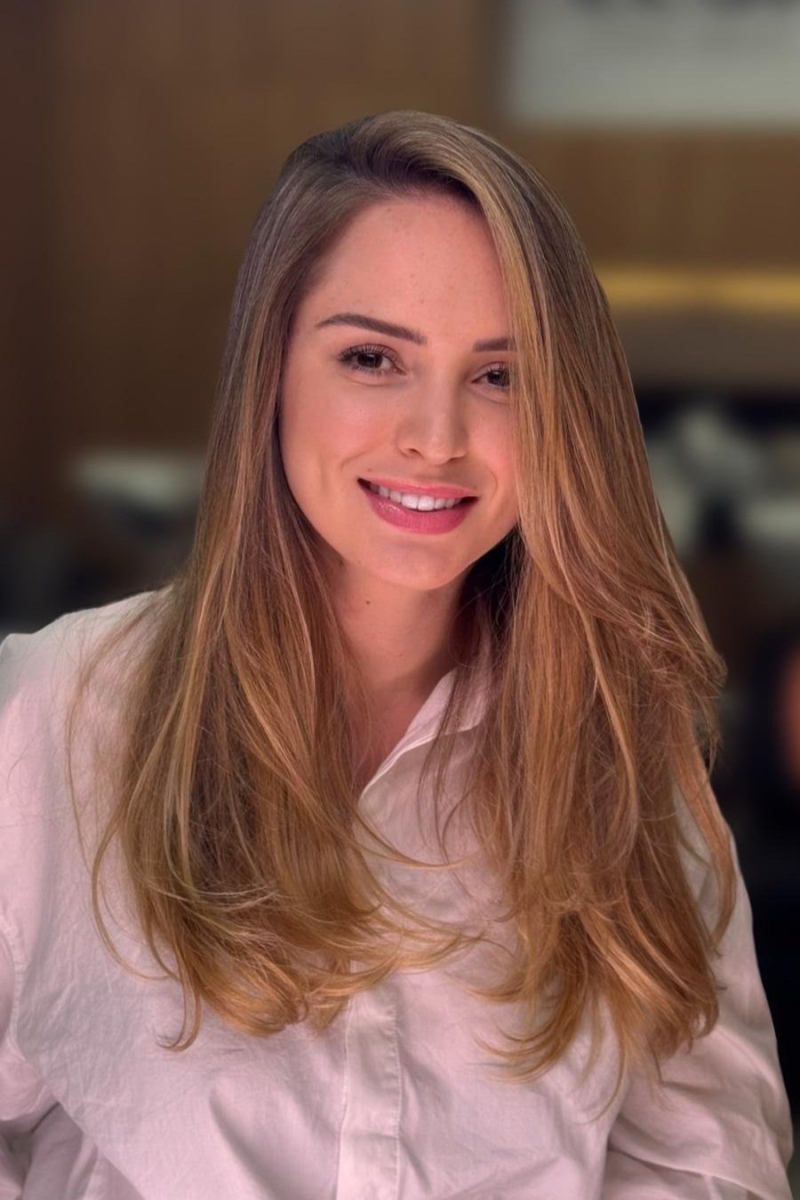 A woman with long, light brown hair smiling, wearing a white shirt, in an indoor setting.