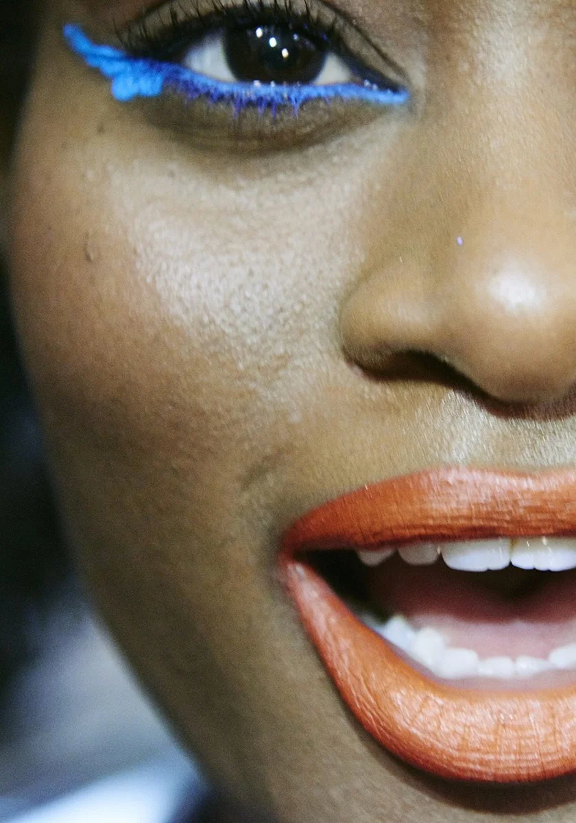 Close-up of a smiling woman's face, showing her eye with blue makeup, nose, and lips with orange lipstick.