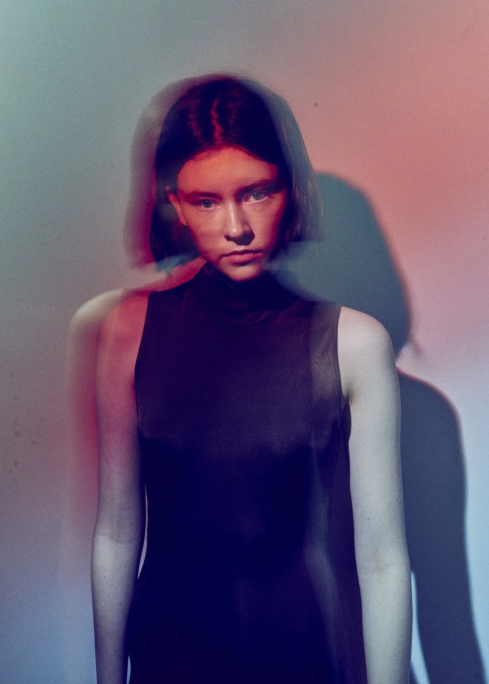 A young woman with short dark hair wearing a sleeveless black top, looking directly at the camera, with colorful lighting creating blue and red shades on her face and background.