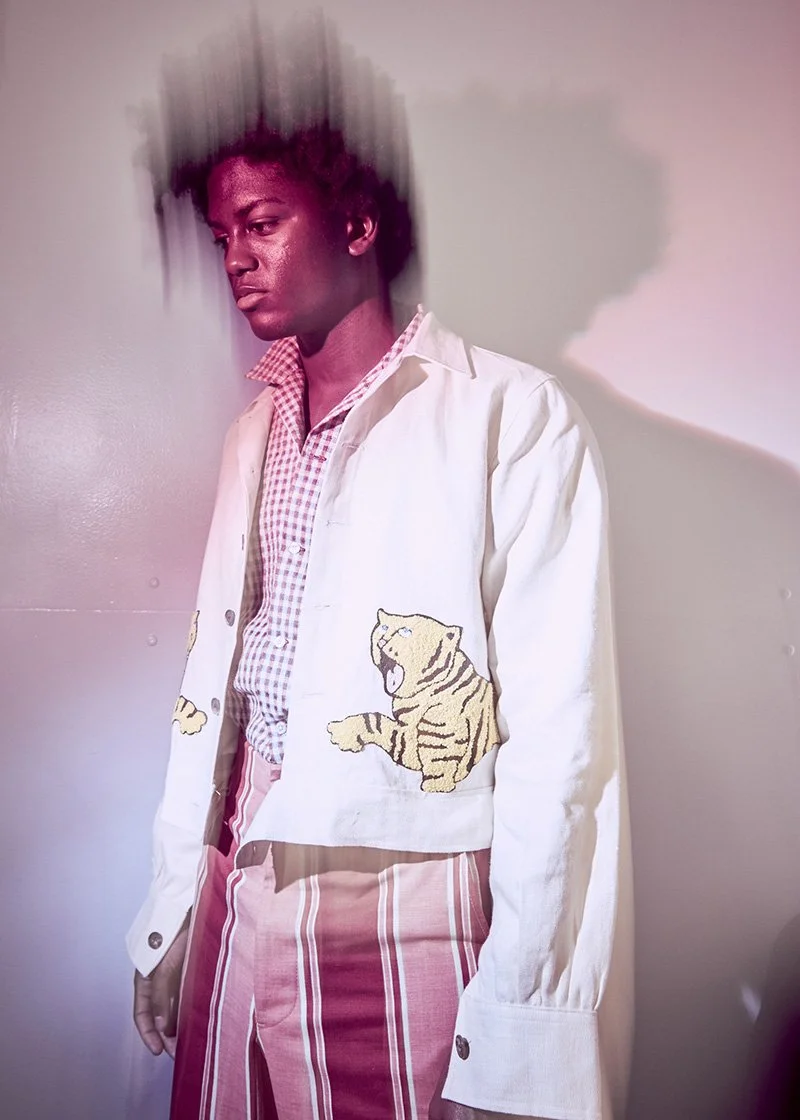 A young person with dark skin and short curly hair, wearing a cream-colored jacket with a tiger patch, a pink checkered shirt, and red and white striped pants, standing against a light-colored wall with a pinkish light and a distortion effect around