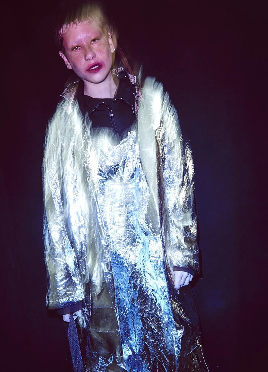 A young person with red hair and freckles wearing a metallic silver and gold jacket over a black zip-up hoodie, standing against a dark background.