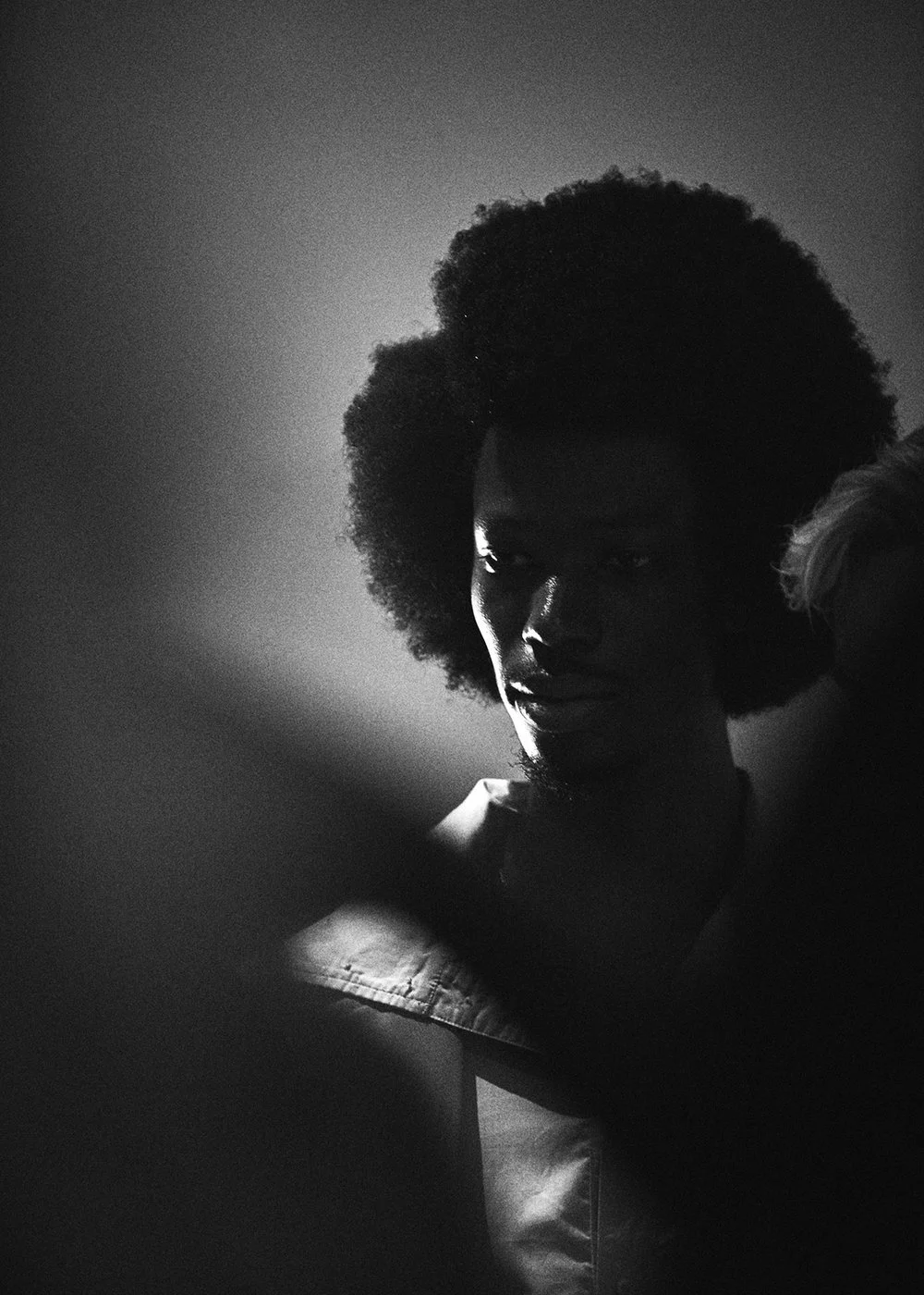 Black and white portrait of a person with natural afro hair, partially illuminated by side lighting, looking down with a pensive expression.