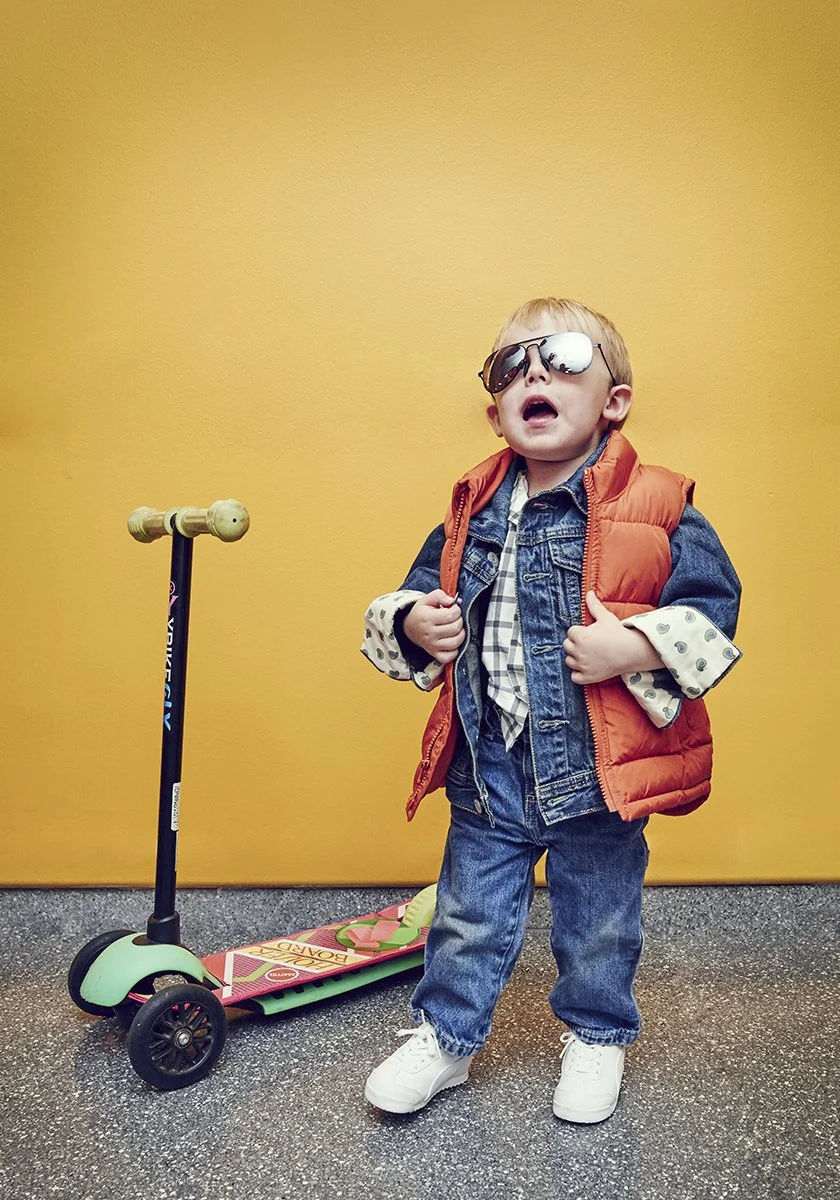 A young boy wearing sunglasses, a denim jacket, and an orange puffy vest, standing with a skateboard beside him against a yellow wall.