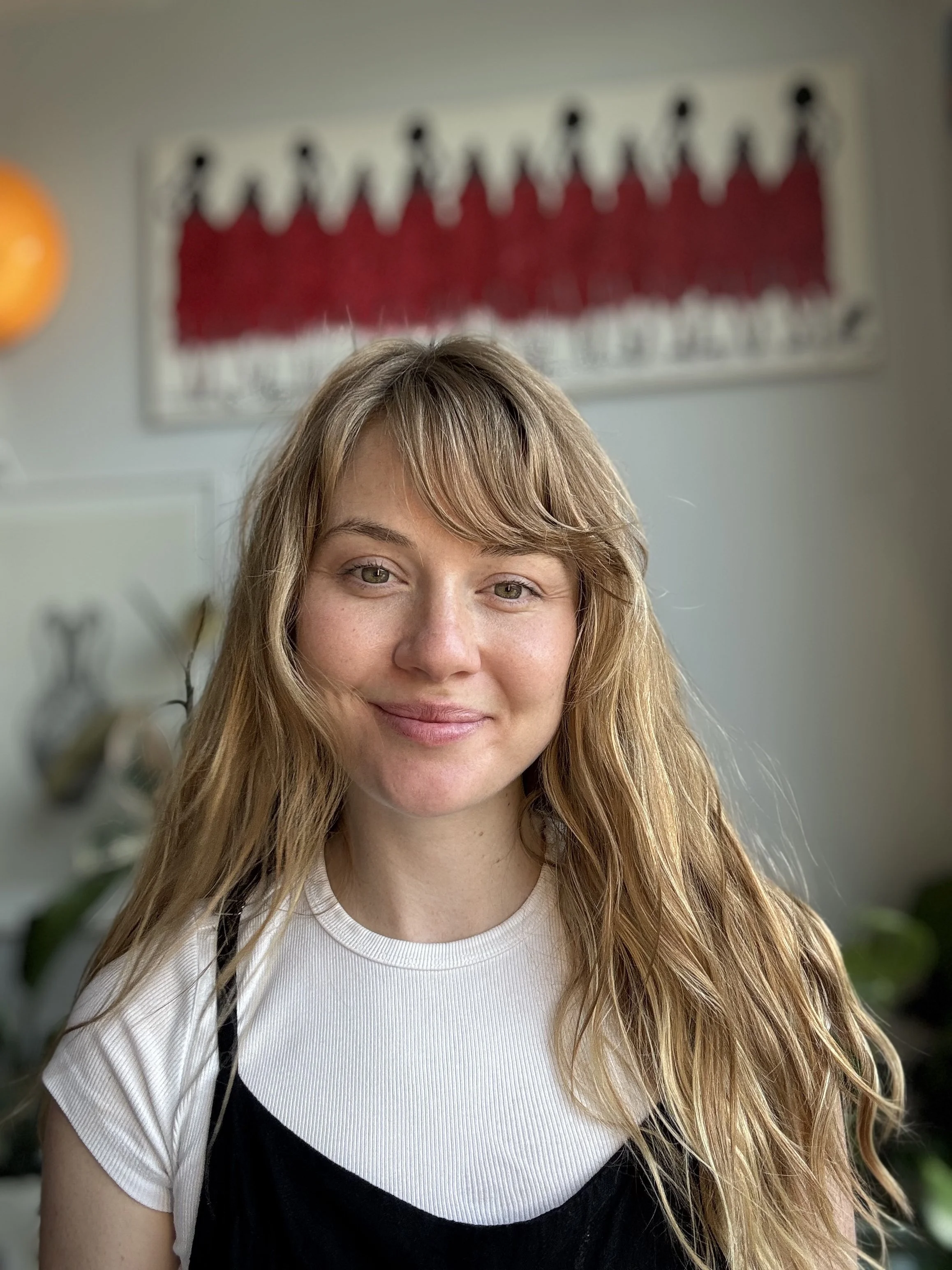 A young woman with long, wavy blonde hair and fair skin, smiling in an indoor setting with a red and white artwork on the wall and some plants in the background.