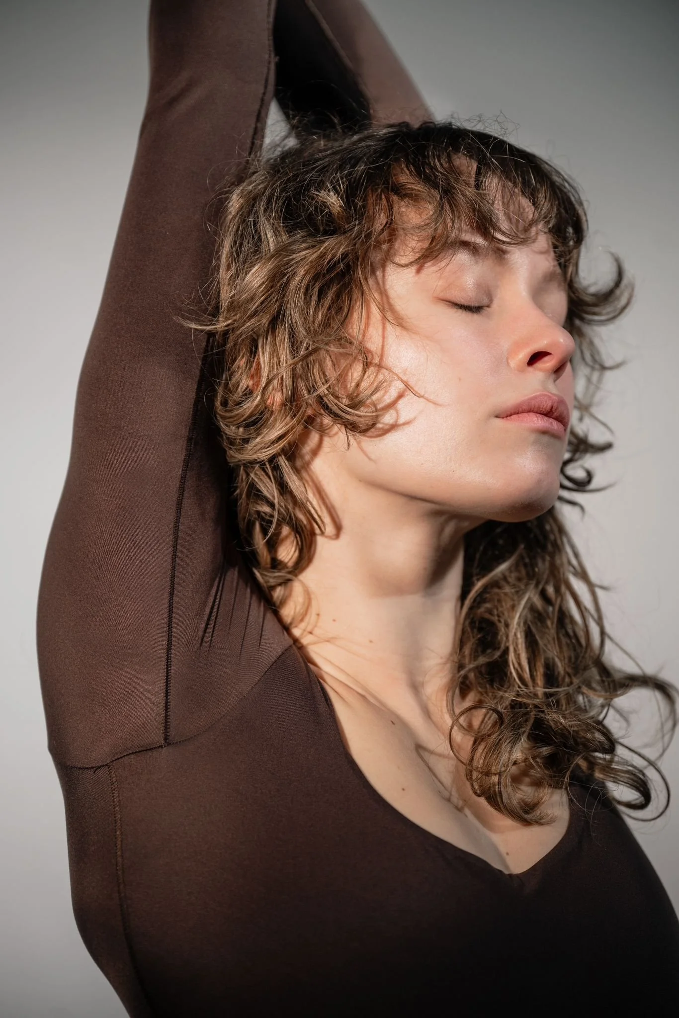 A woman with curly brown hair and fair skin, eyes closed, stretching her arm above her head wearing a brown long-sleeve shirt, against a plain gray background.