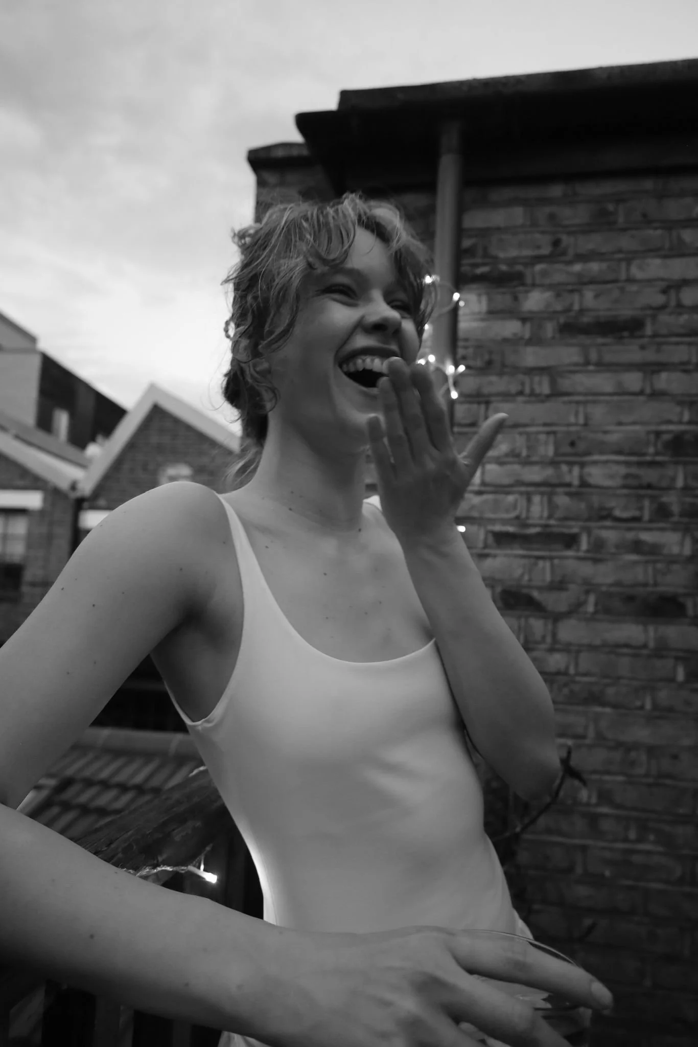 A woman with short wavy hair laughing, covering her mouth with her hand, standing outside with brick and house rooftops in the background.
