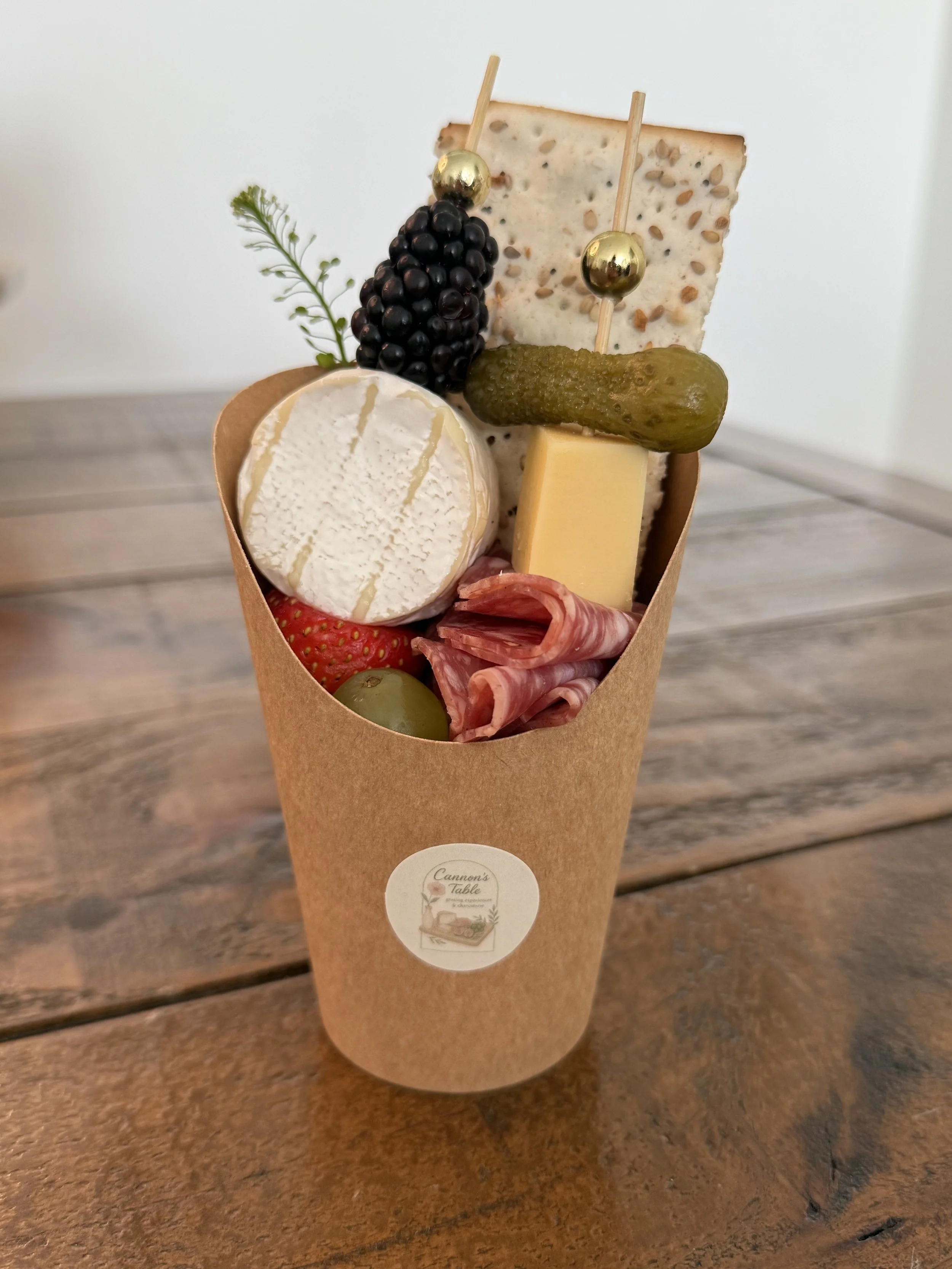 Gourmet charcuterie cup with cheese, cured meat, grapes, strawberry, a pickle, and crackers created by Cannon's Table.
