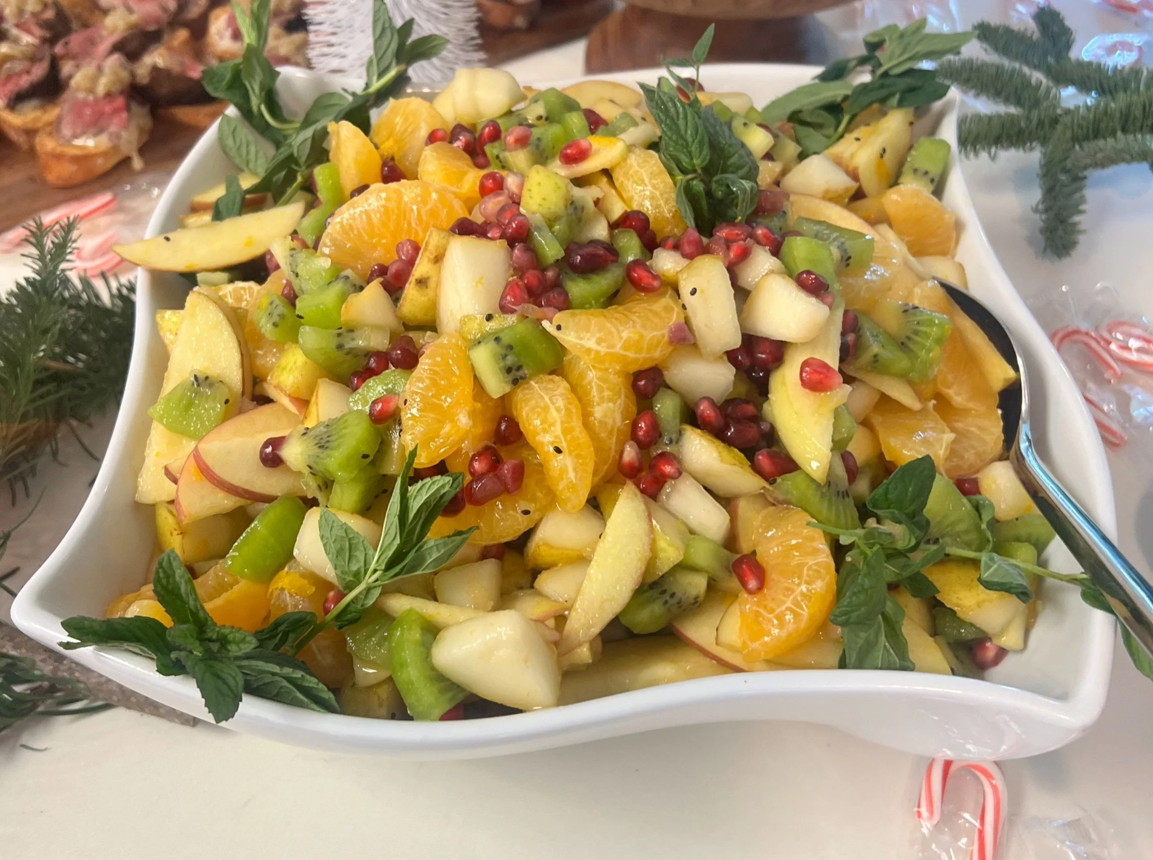 A bowl of mixed fruit salad with oranges, apples, kiwi, pomegranate seeds, and mint leaves created by Cannon's Table.