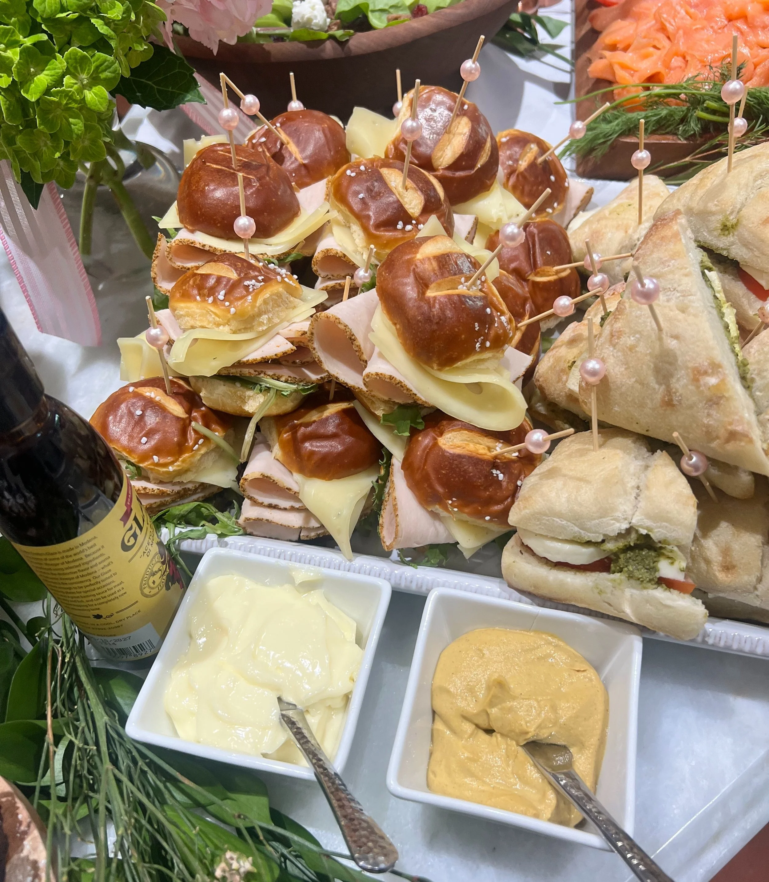 Assorted appetizers including mini sandwiches with cheese, deli meat, and pickles, with toothpicks, alongside bowls of mayonnaise and mustard, on a white platter with green herbs and flowers for garnish created by Cannon's Table.