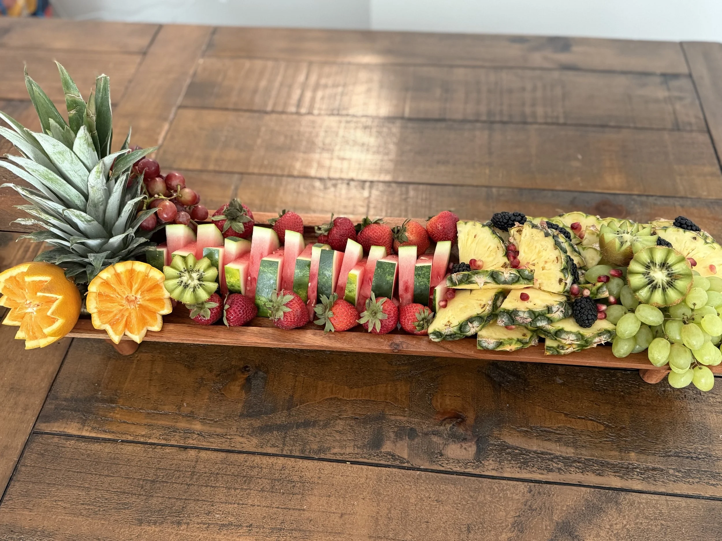 A long wooden platter with various sliced and whole tropical fruits including pineapple, watermelon, strawberries, kiwi, green grapes, blackberries, and red grapes created by Cannon's Table.