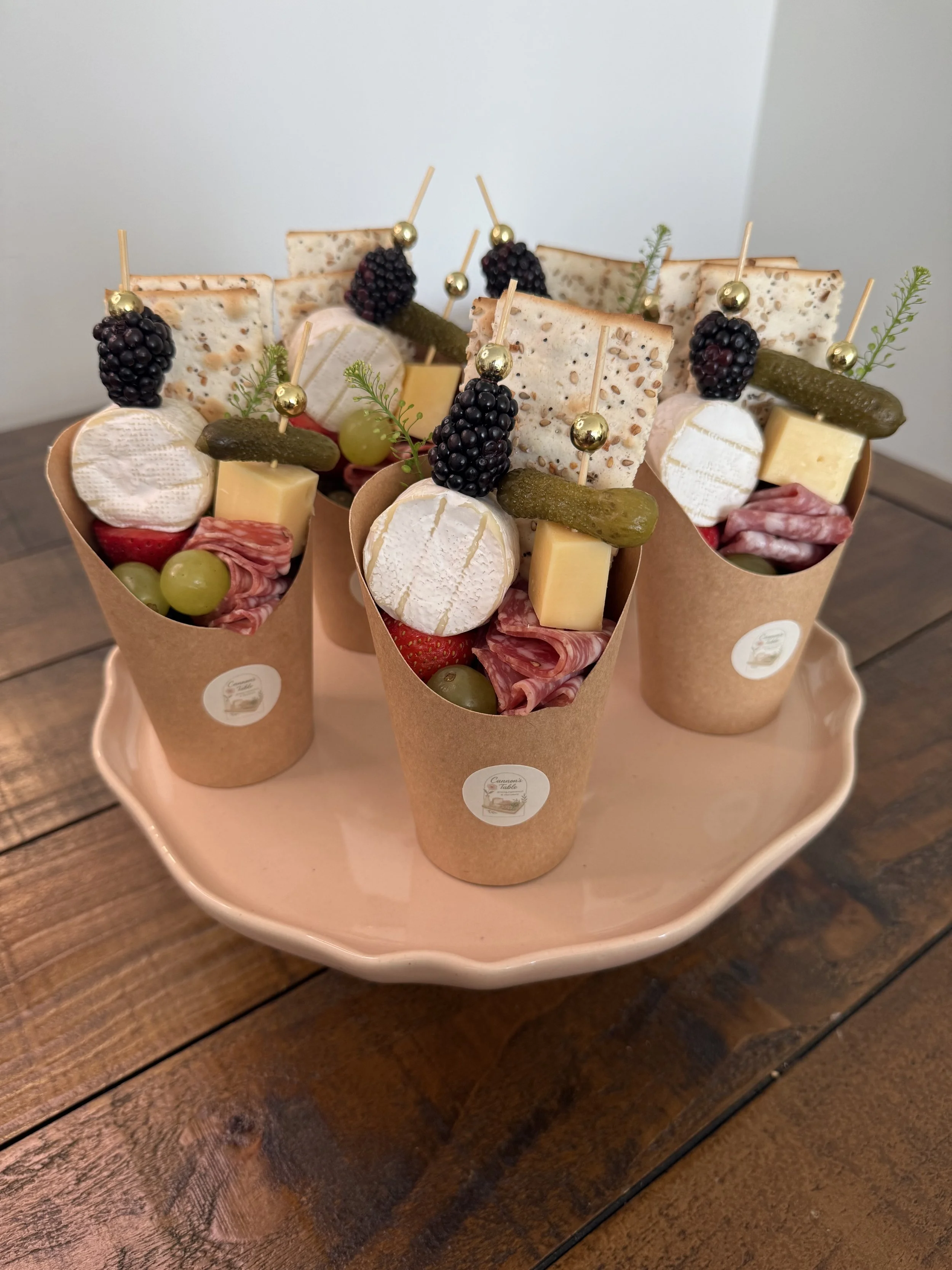 Charcuterie cups with cheese, crackers, blackberries, grapes, pickles, cured meats, and herbs, arranged on a pink plate created by Cannon's Table.