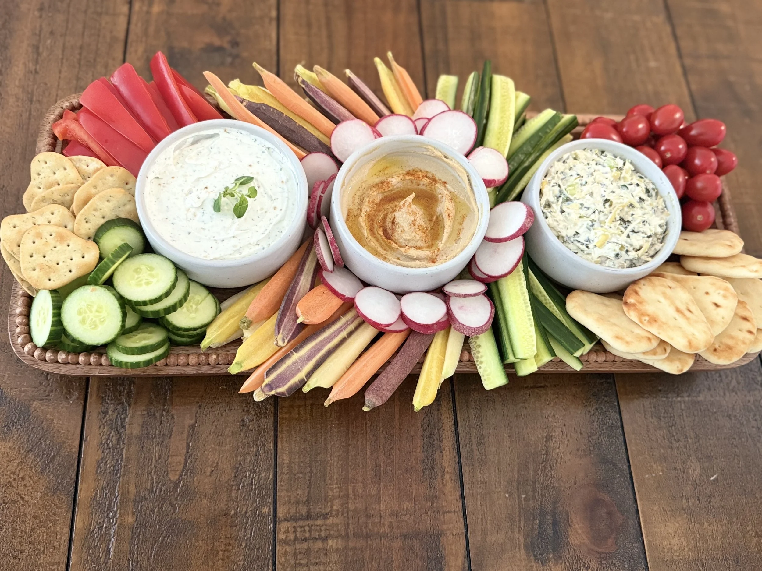 A vegetable platter with three dips accompanied by sliced cucumbers, cherry tomatoes, radishes, crackers, and assorted colorful vegetable sticks on a wooden tray created by Cannon's Table.