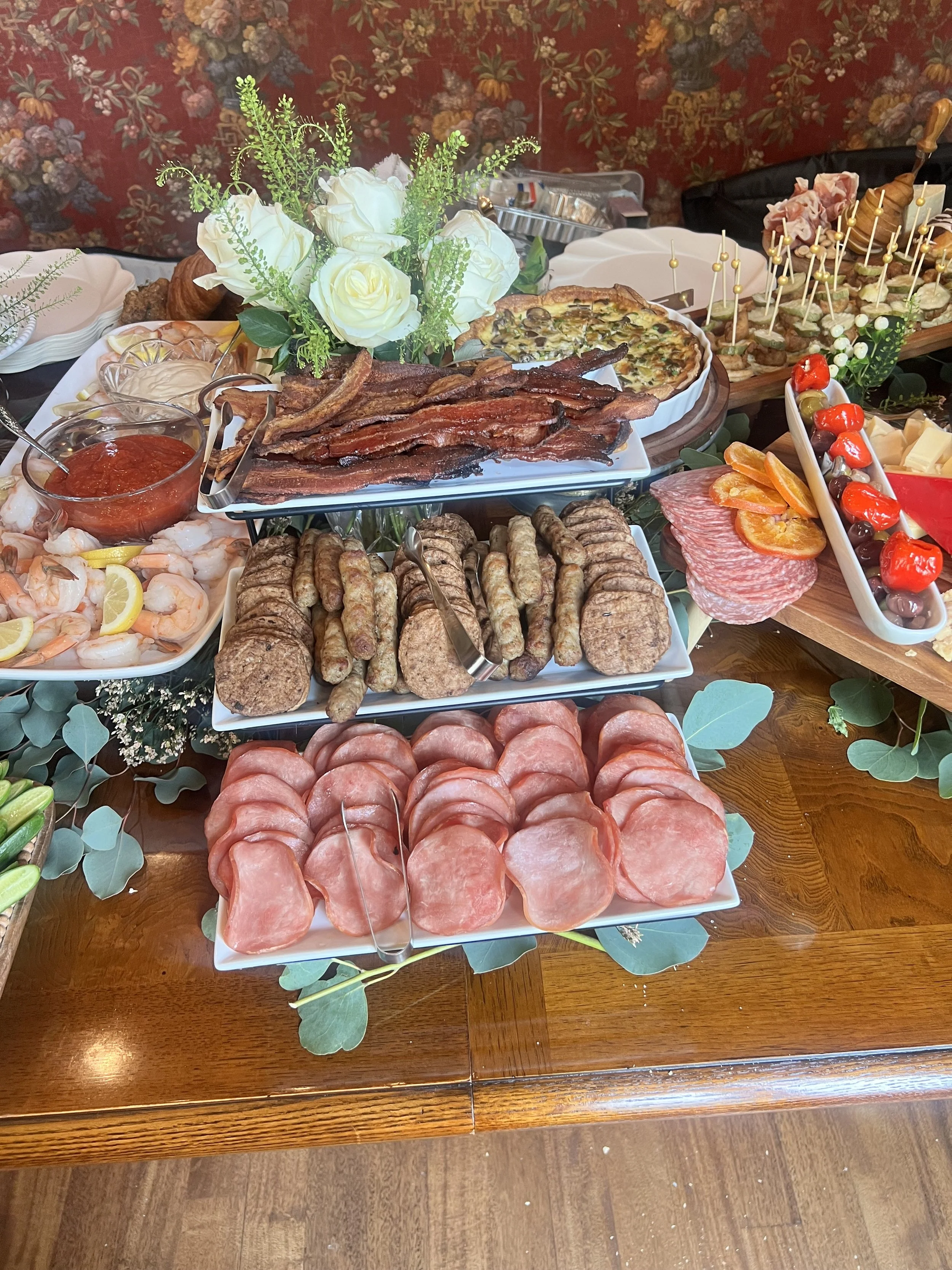 A grazing table with various meats, seafood, cheeses, and appetizers, decorated with green leaves and white roses created by Cannon's Table.