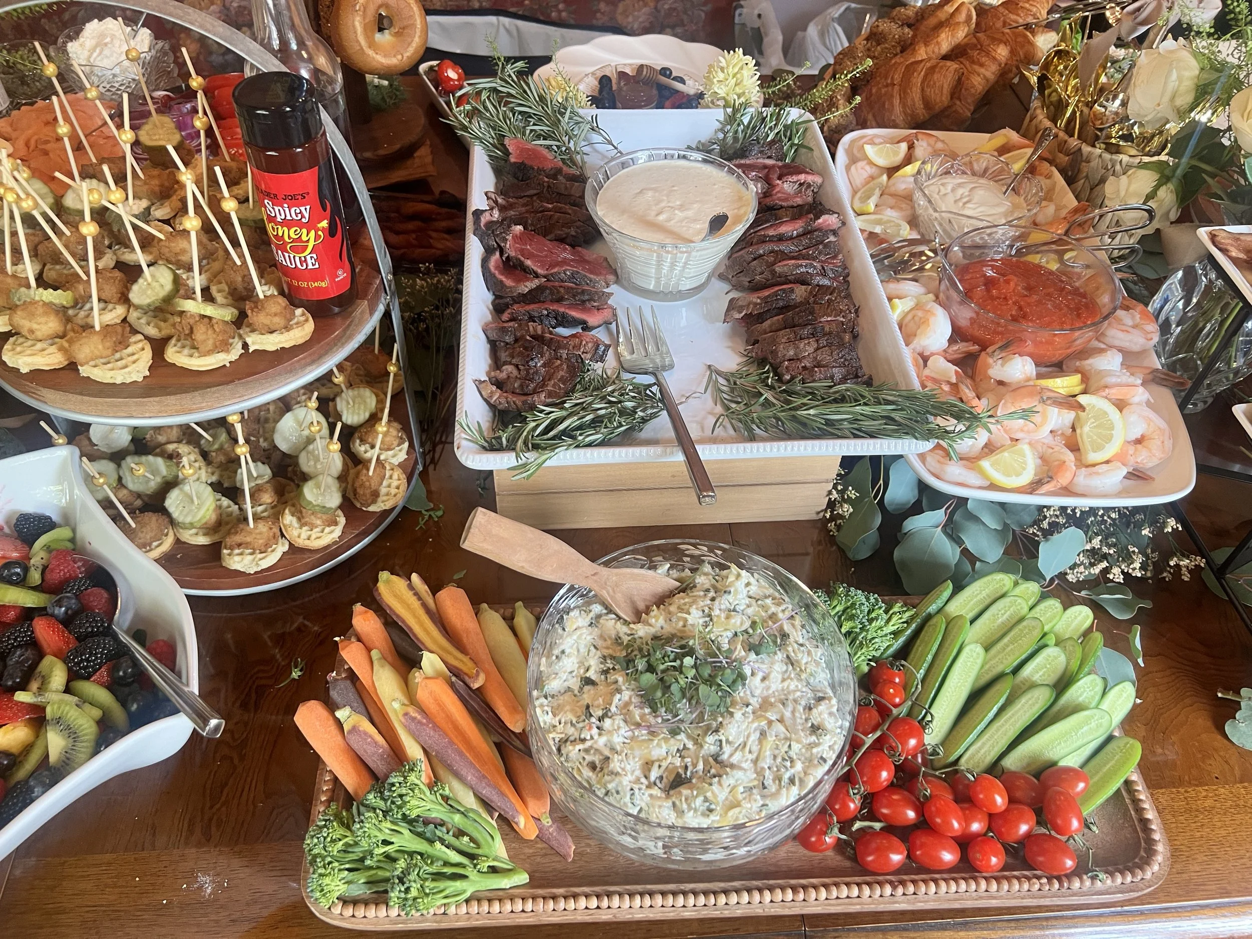 A variety of appetizers and finger foods on a grazing table, including fresh vegetables, shrimp, sliced steak with dipping sauce, fruit skewers, and a bowl of creamy salad created by Cannon's Table.